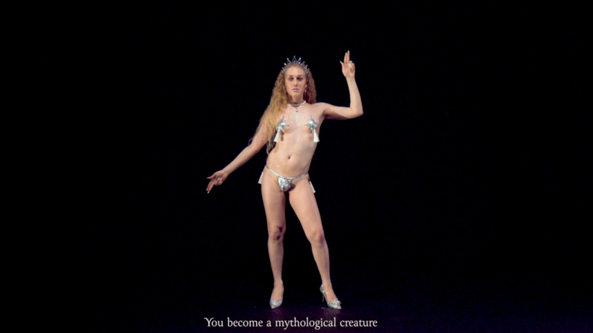 A person with long blonde hair is wearing a tiara, thong, and heels while dancing in front of a black background.