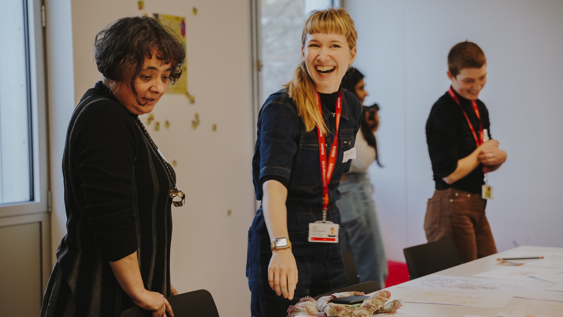 Image shows two people laughing in an art classroom. Ruby has blonde hair and is wearing a denim boiler suit.