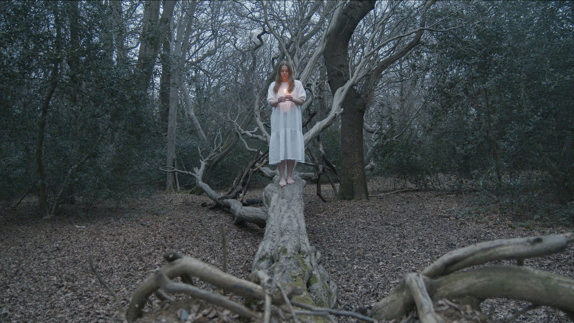 A woman wearing a white dress is looking down at her hands holding a candle and is standing on a fallen tree in the woods