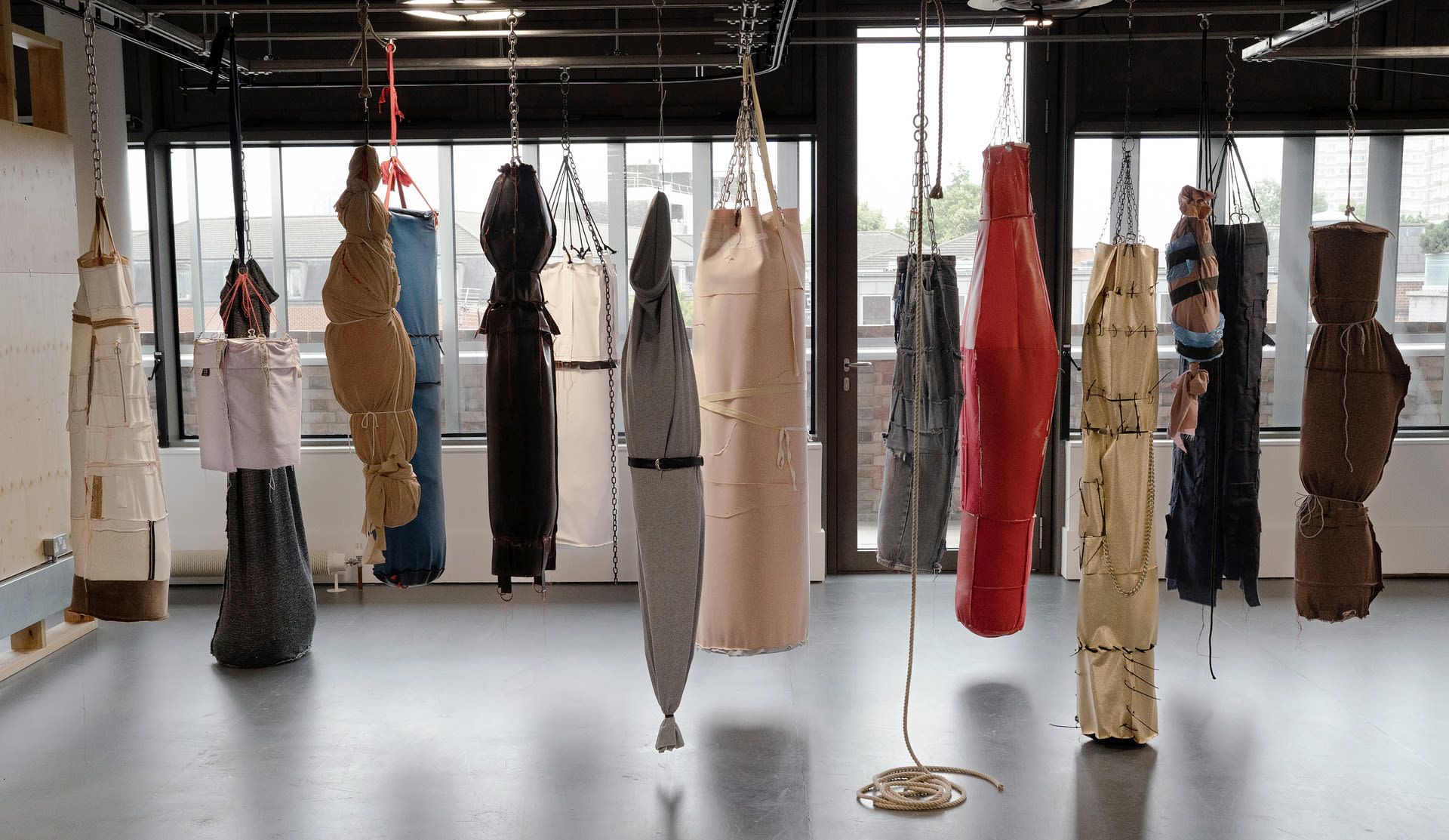 A series of 15 objects made from various fabrics hang from metal chains, resembling punch bags