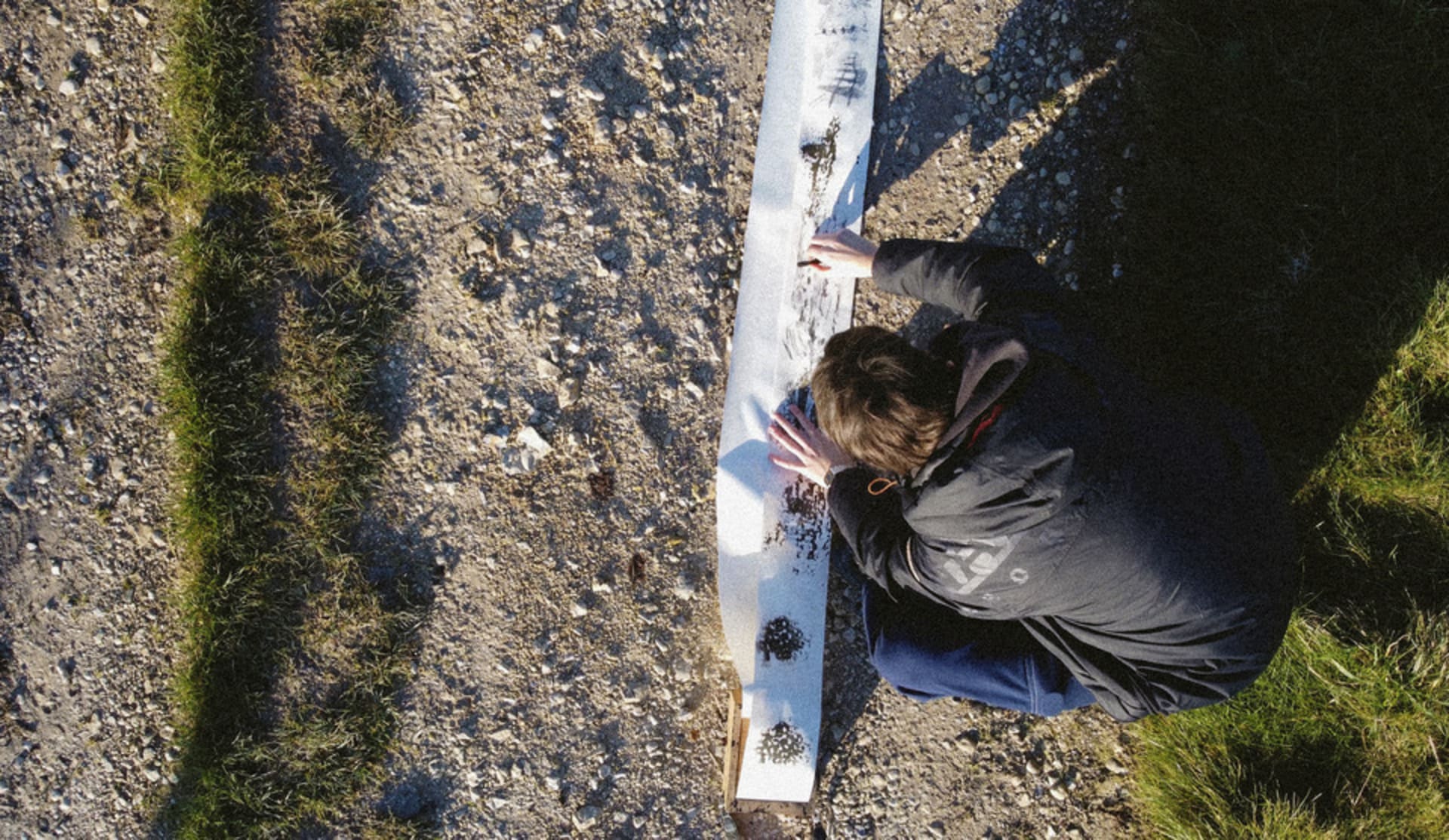 Picture is an arial photograph showing a long scroll of paper on a dirt track. A figure is crouched over the paper making marks.