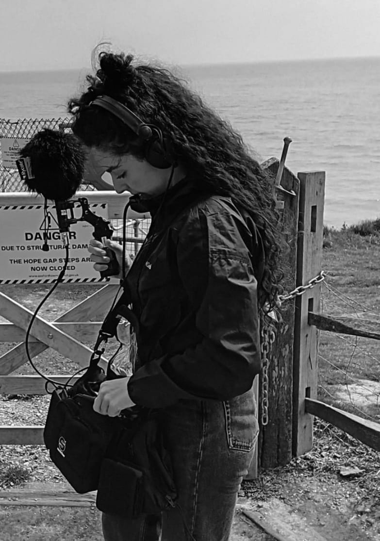 A photograph of a woman with long, dark, curly hair by the sea. She is holding a microphone and wearing headphones. 