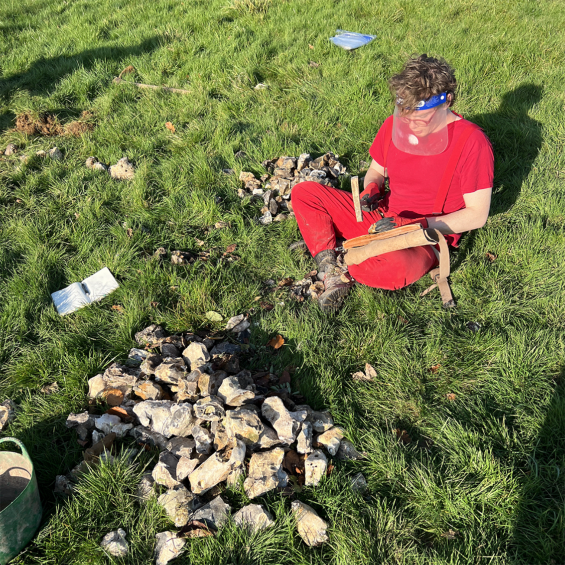 Quentin wearing red sitting on the ground in a field knapping flint 