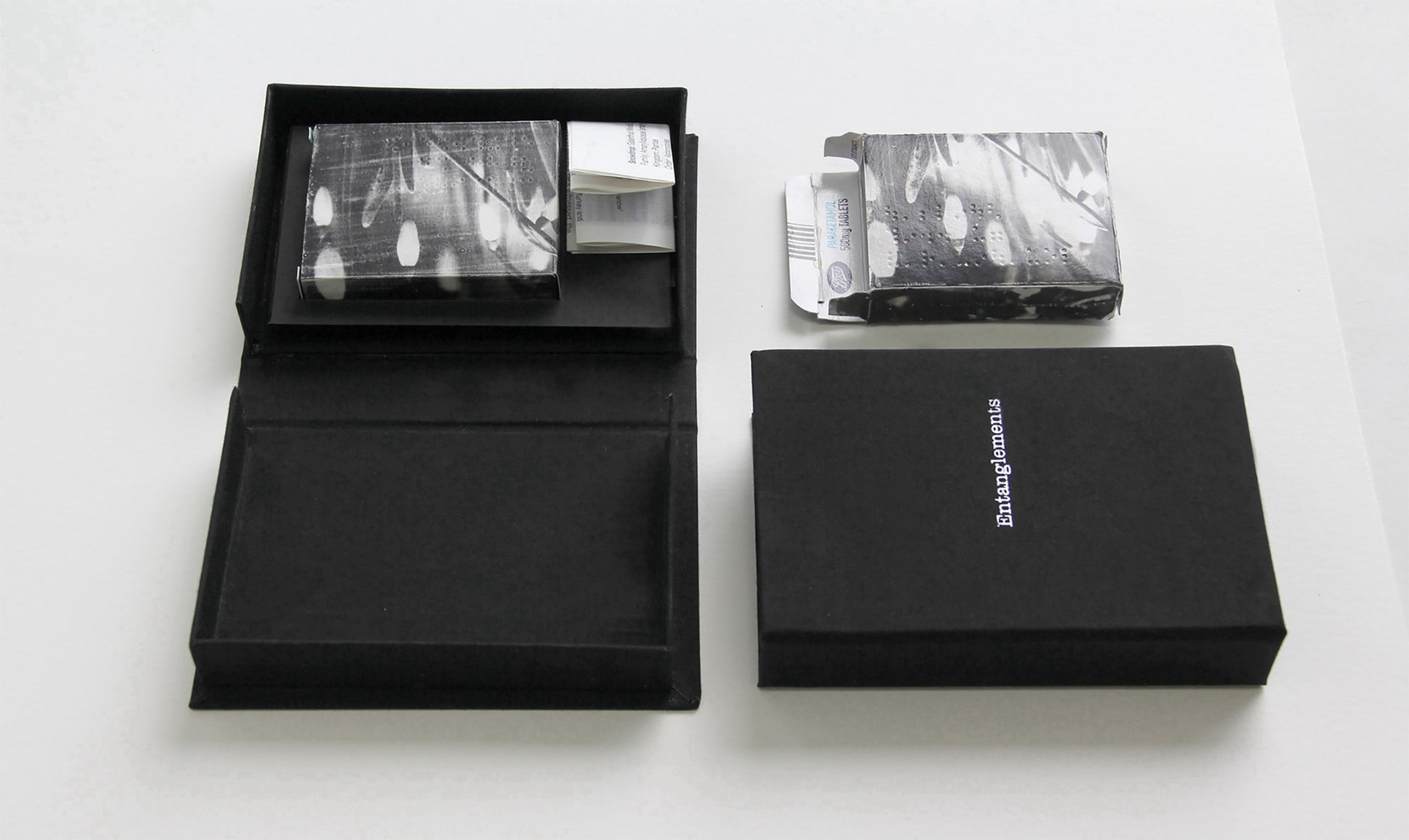 two black clamshell boxes containing recycled paracetamol boxes printed with a snowdrop image, one is open