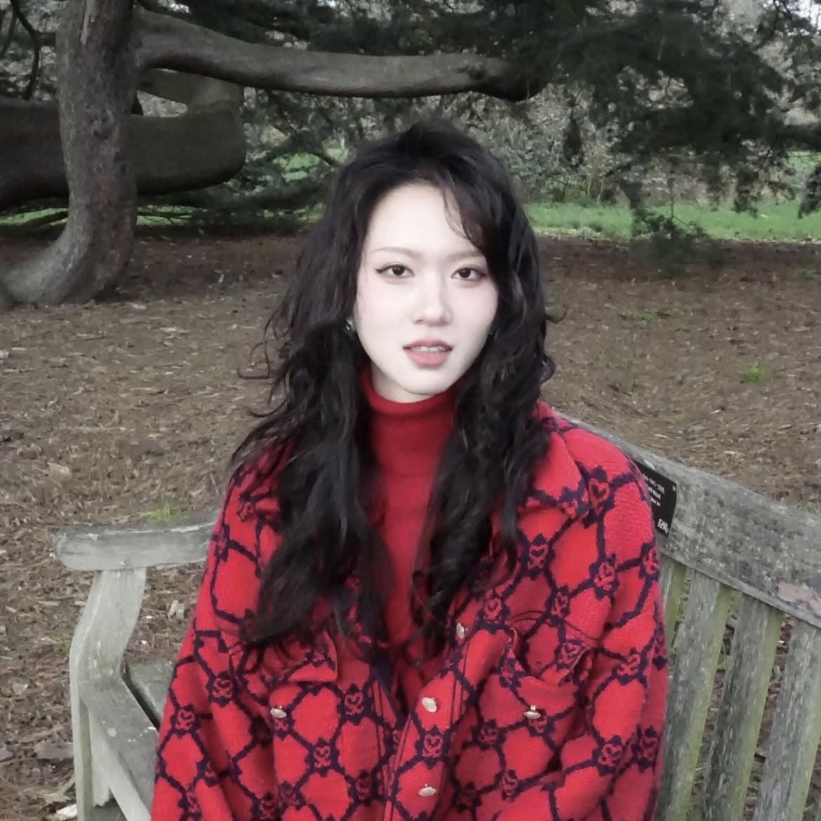 A girl with long curly hair wearing a red jacket, the background is in a park with trees and grass.