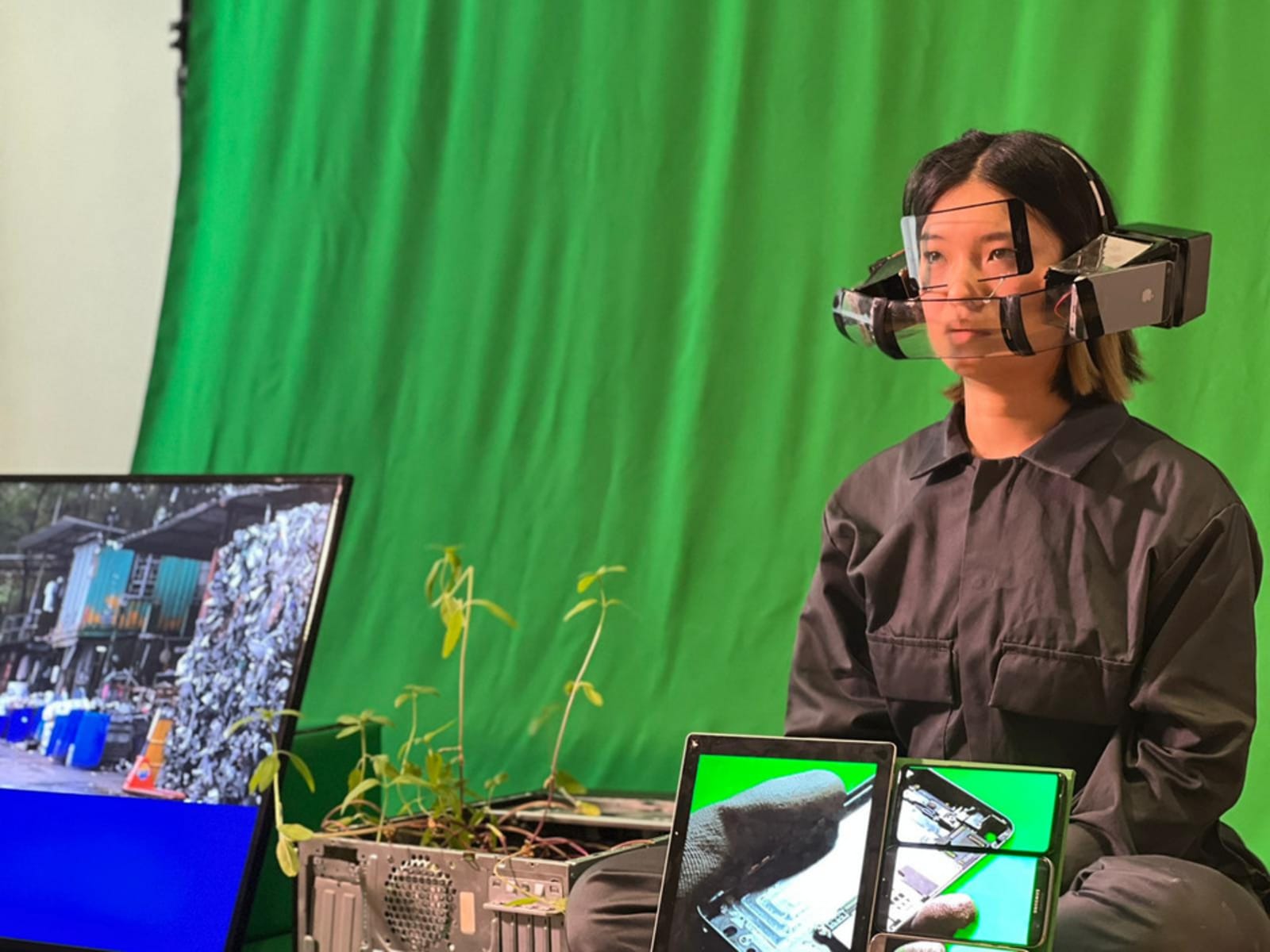 Image of student, wearing boiler suit and safety mask made out of salvaged technology, in studio setup with greenscreen backdrop