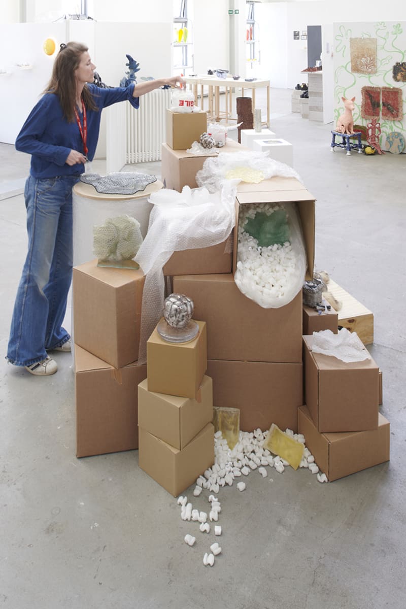 a woman in blue trousers reaches across a pile of cardboard boxes that are plinths to various works in glass 