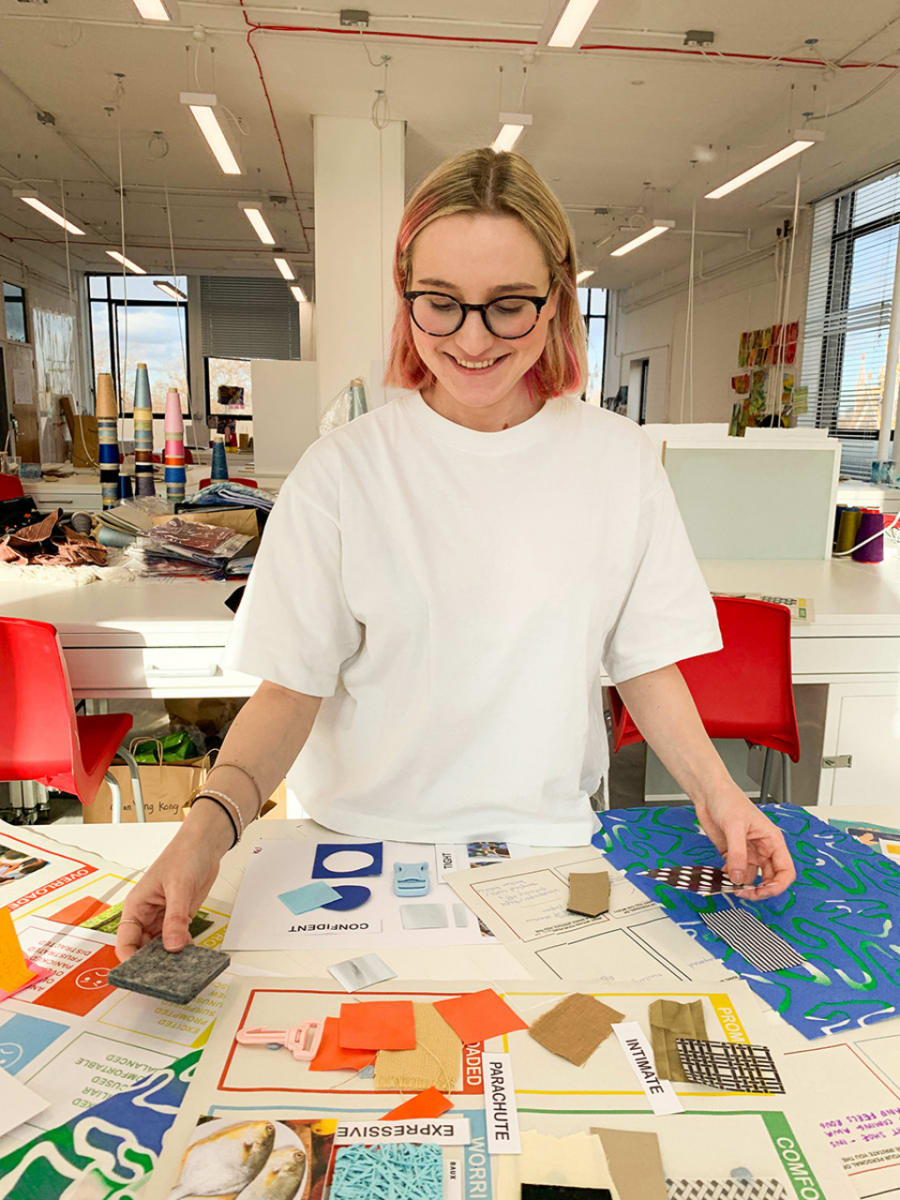 Young female, in white t-shirt and glasses, short blonde-pink hair,  working by the table full of fabric and paper swatches.