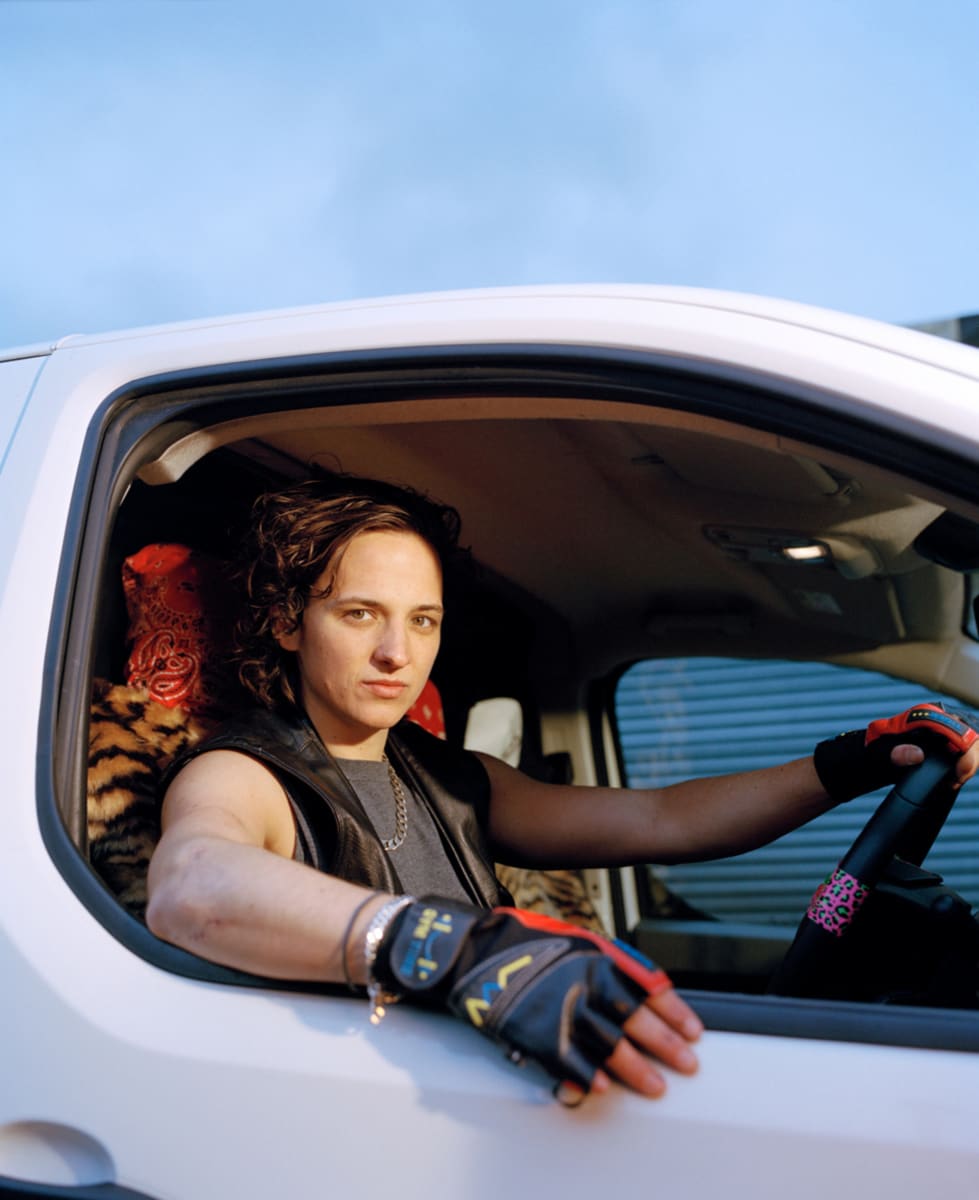 Photograph of a dyke sitting behind the wheel of a white van, wearing gym gloves. The van seat is leopard print fabric