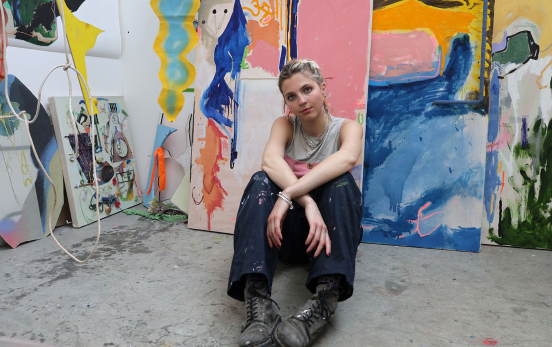 Image of artist sitting on the floor in their RCA studio surrounded by her paintings and sculptures.