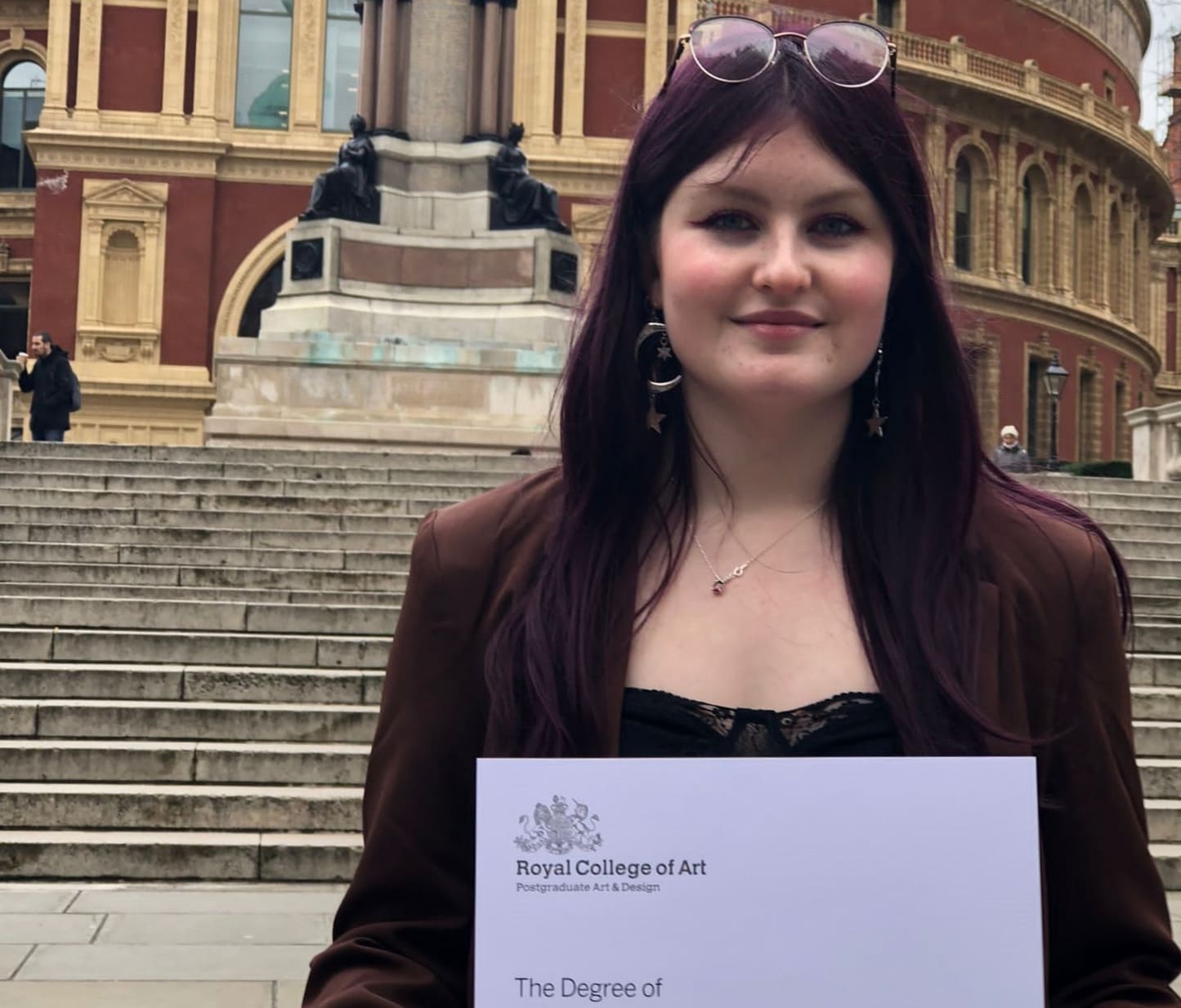 Woman (Lucy Roche) standing in front of the Royal Albert Hall holding a degree certificate after the graduation ceremony.