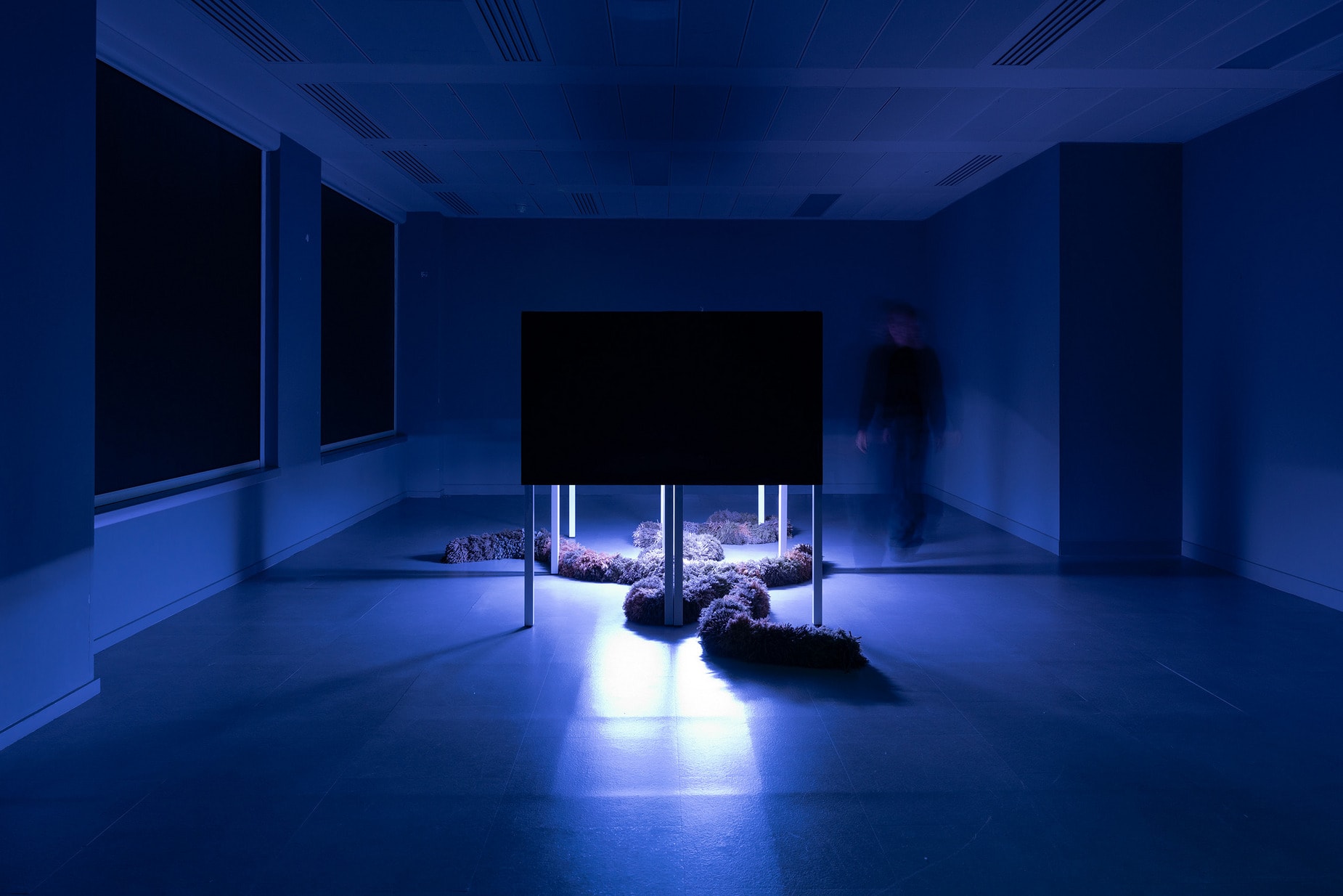 Dark exhibition room with a purple light source, illuminating a central installation box, surrounded by textile art pieces.