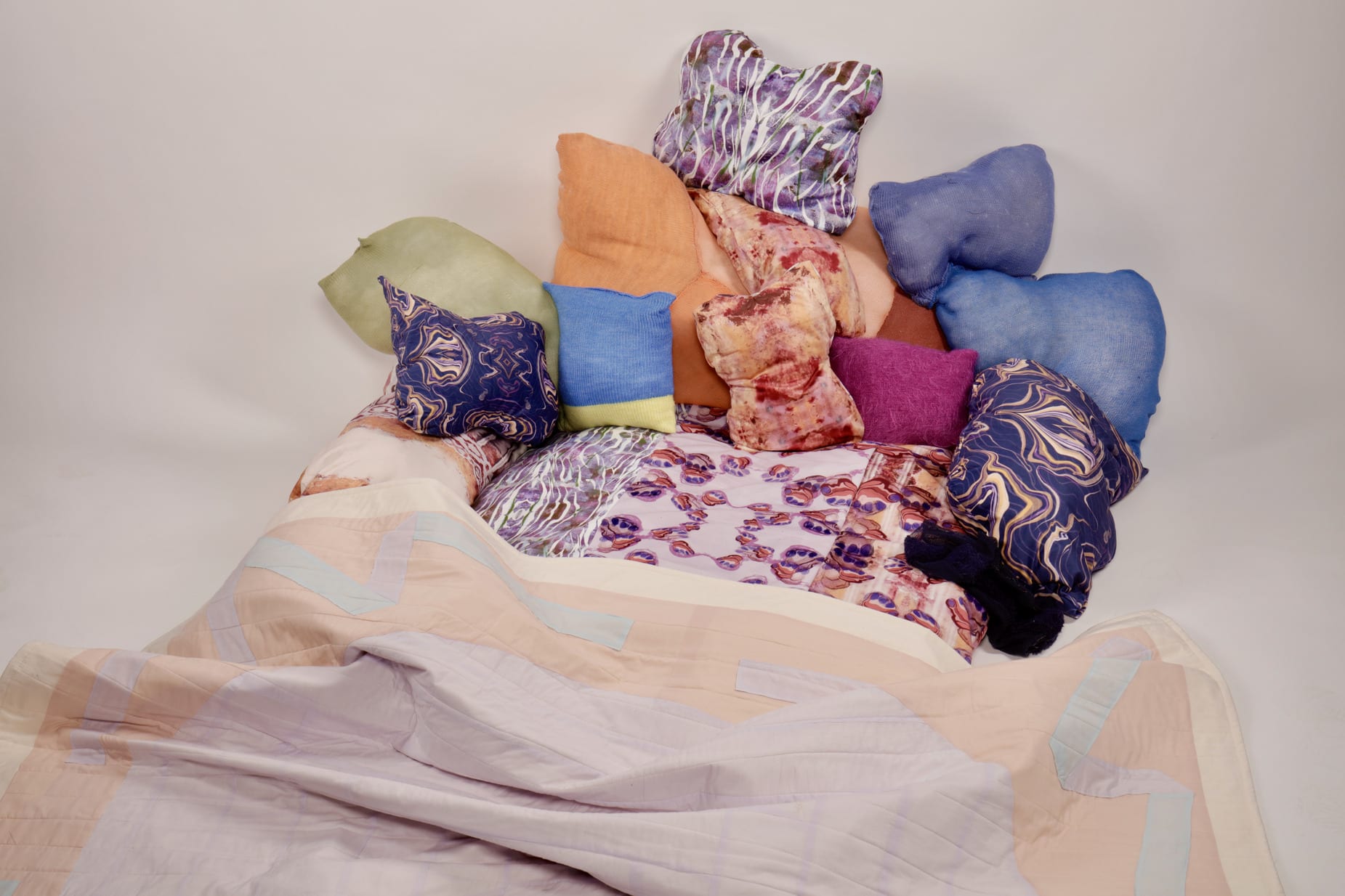 A large cushion with an abstract pattern covered with knitted and abstract print pillows and a pastel coloured quilt.