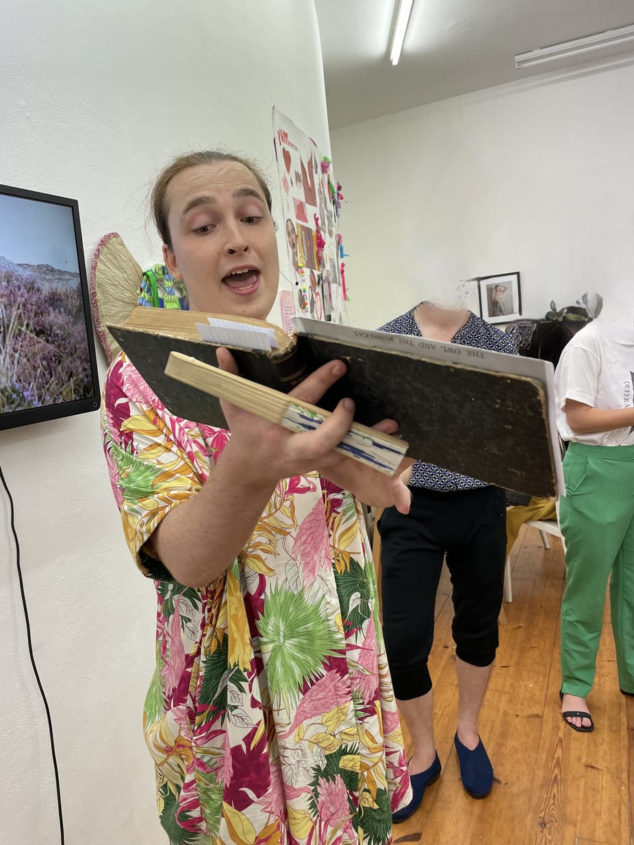 Cas is wearing a floral shirt, holding an old book and speaking at an exhibition they curated. The audiences' faces are blurred.