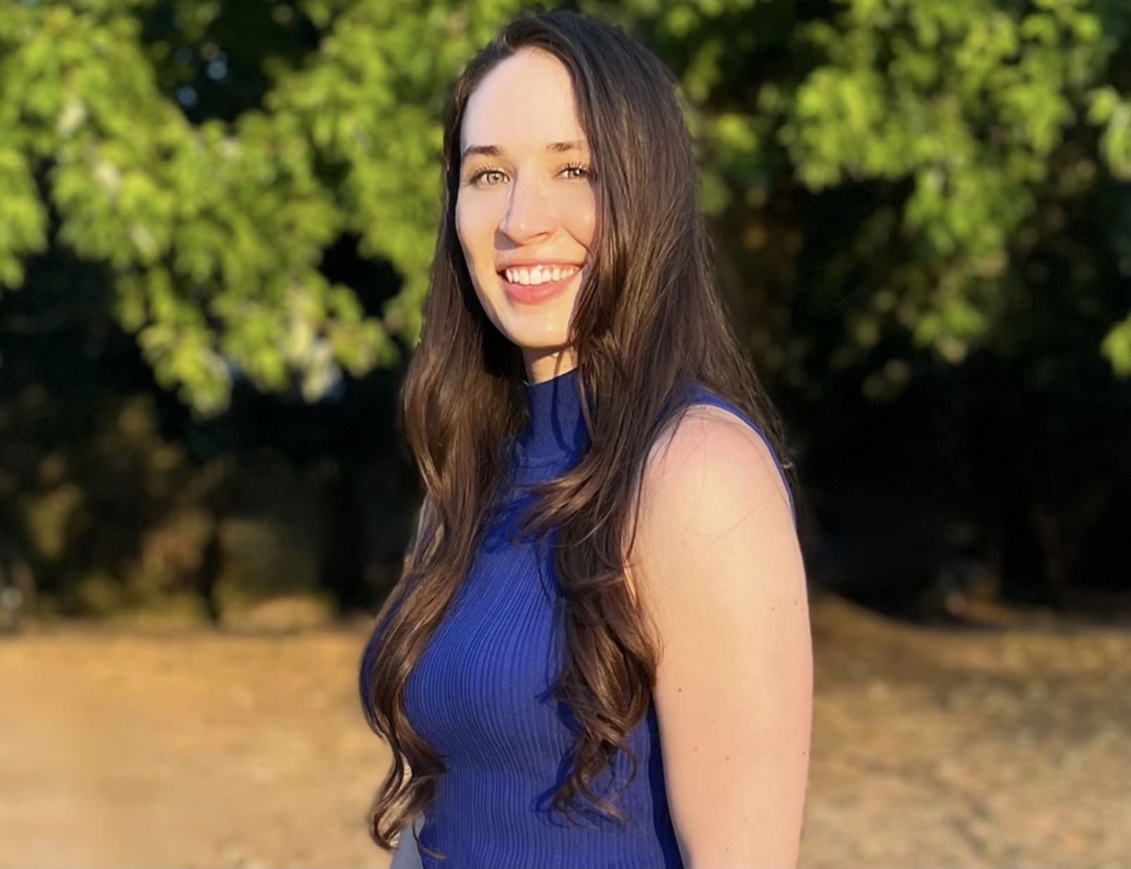 A picture of a woman from the waist up standing in a park, with long brown hair and a blue top looking at the camera.