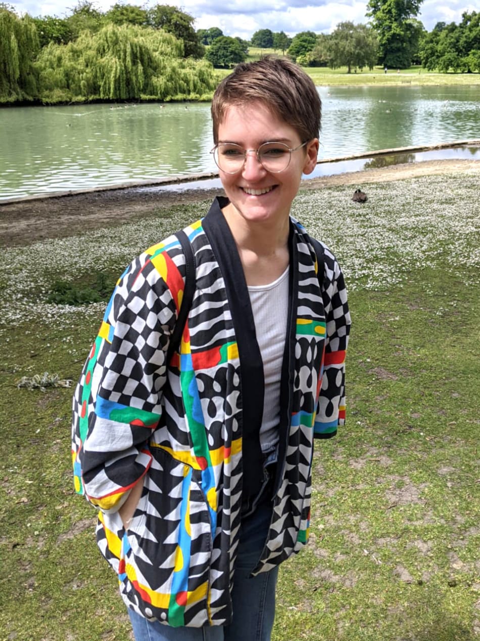 Sarah standing in front of a lake, wearing her classic rainbow patterned kimono jacket and smiling towards the bottom left.