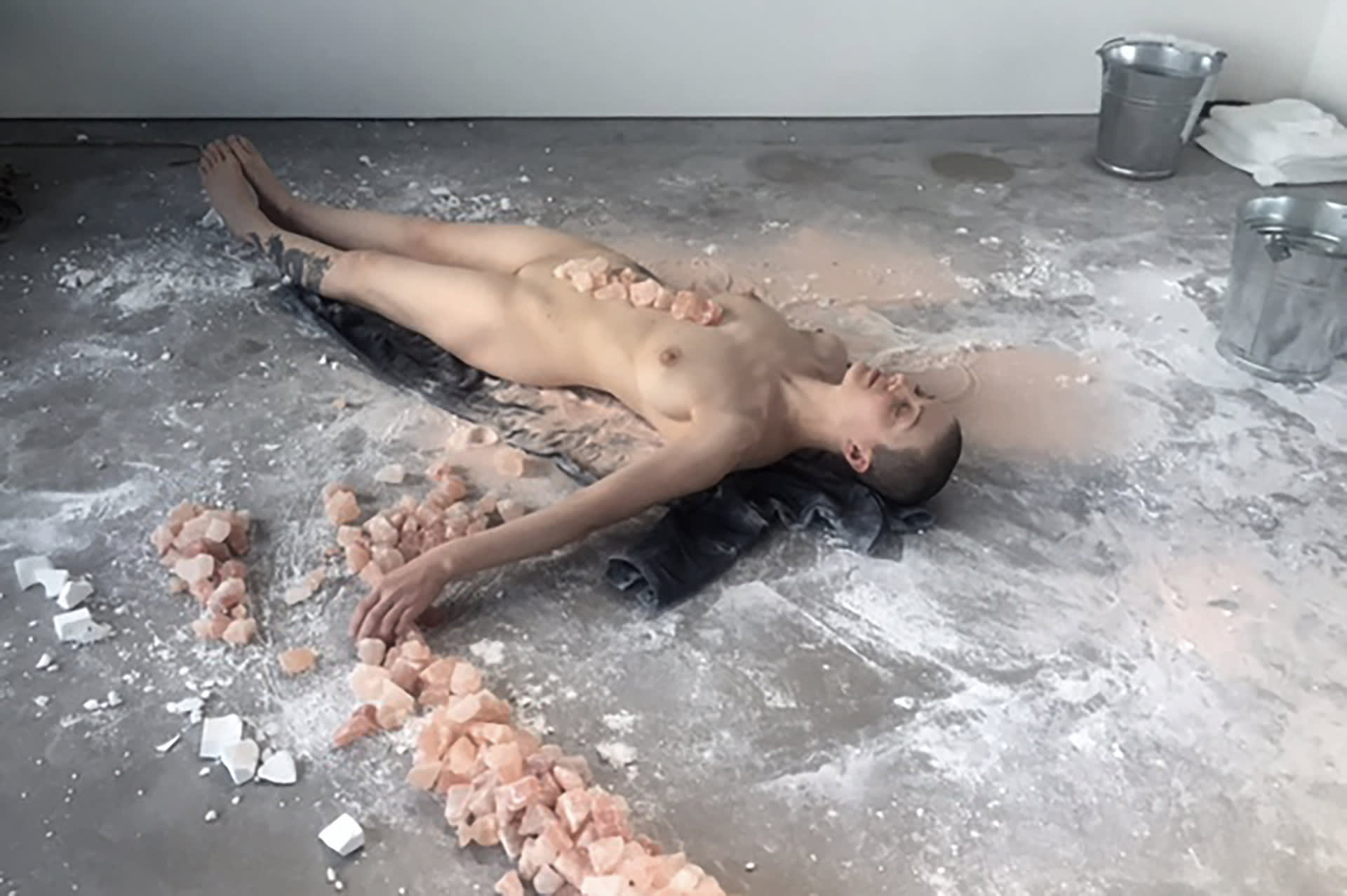 Naked, shaved-head, female performer lying on the floor with Himalayan salt rocks on her torso, aligned to her spine. 