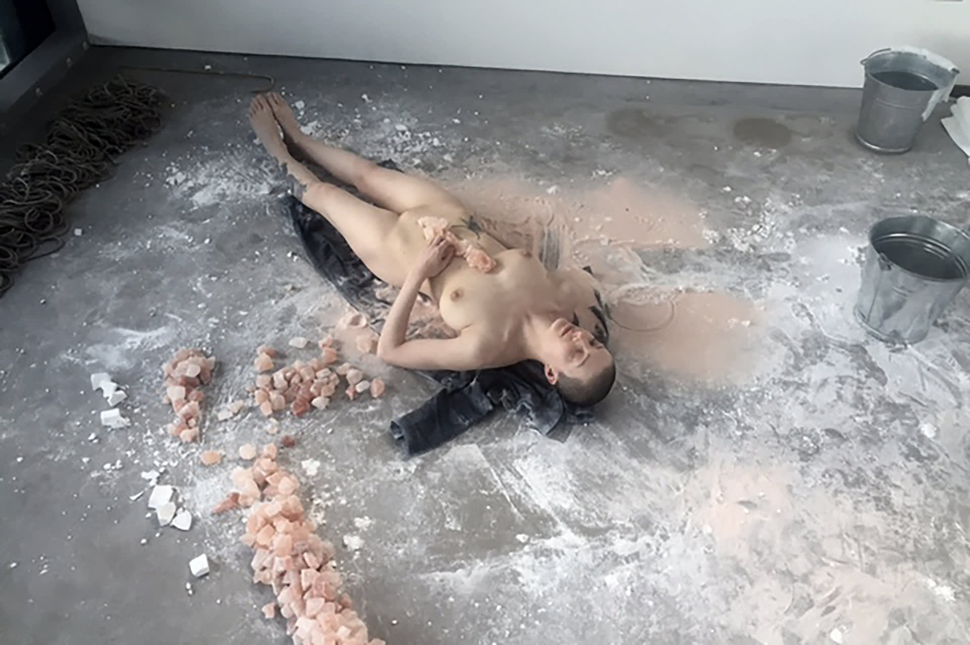 Naked, shaved-head, female performer lying on the floor with Himalayan salt rocks on her torso, aligned to her spine. 