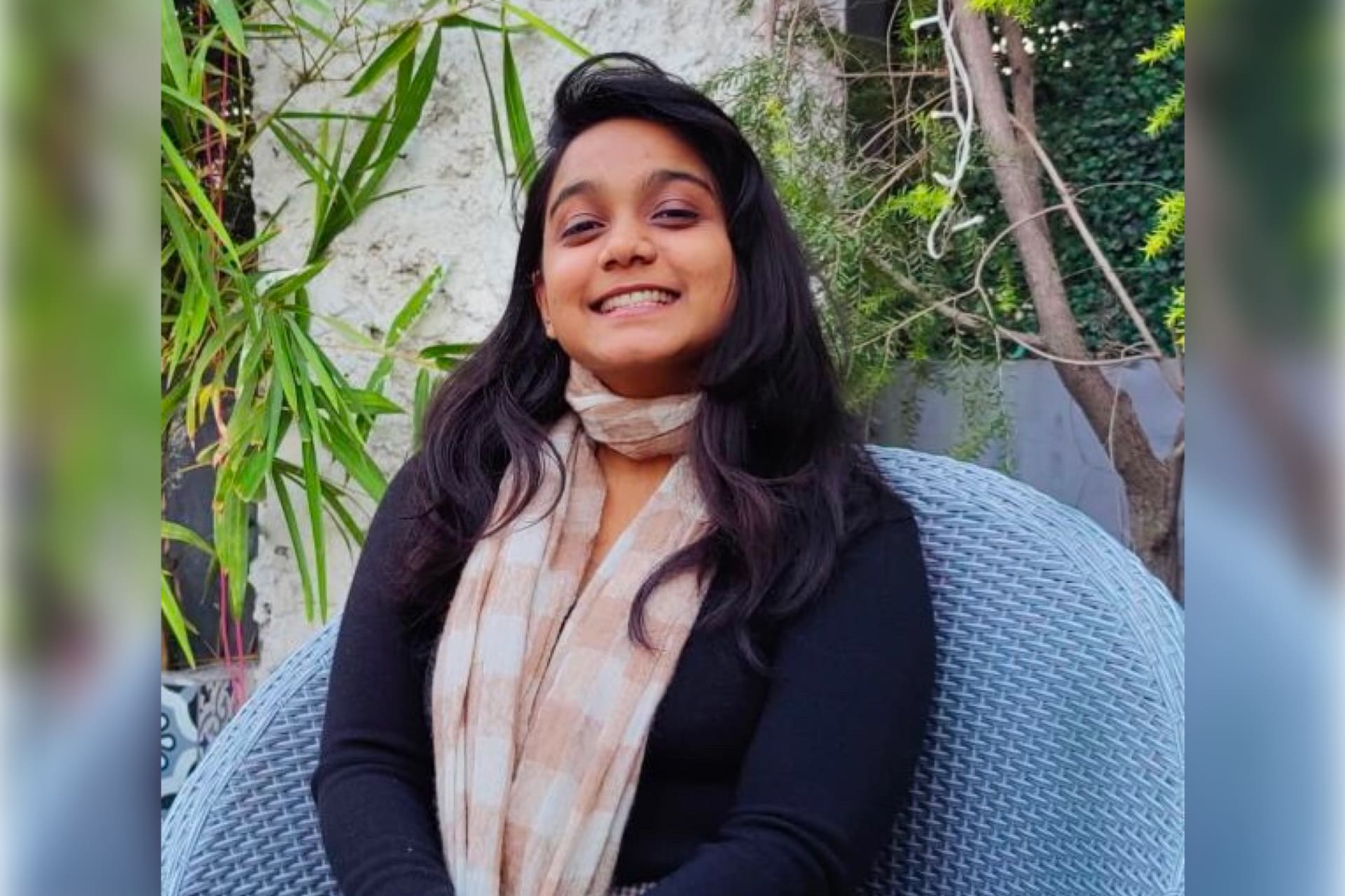 A happy-looking Indian female in a black sweater and a beige checked scarf, sitting on a blue garden chair among plants