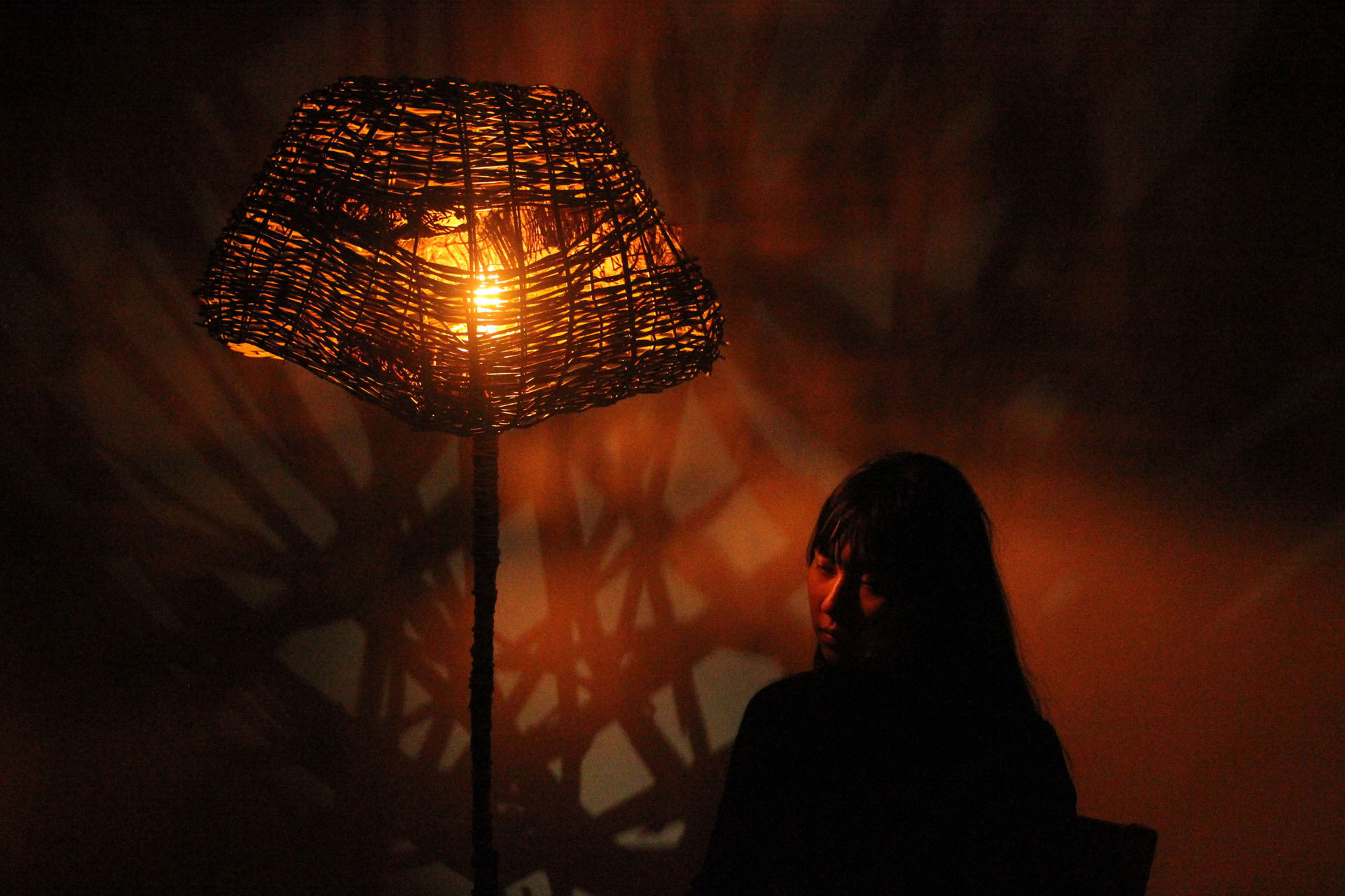Willow woven lampshade lit from within throwing irregular shadows on wall behind with girl sat below right