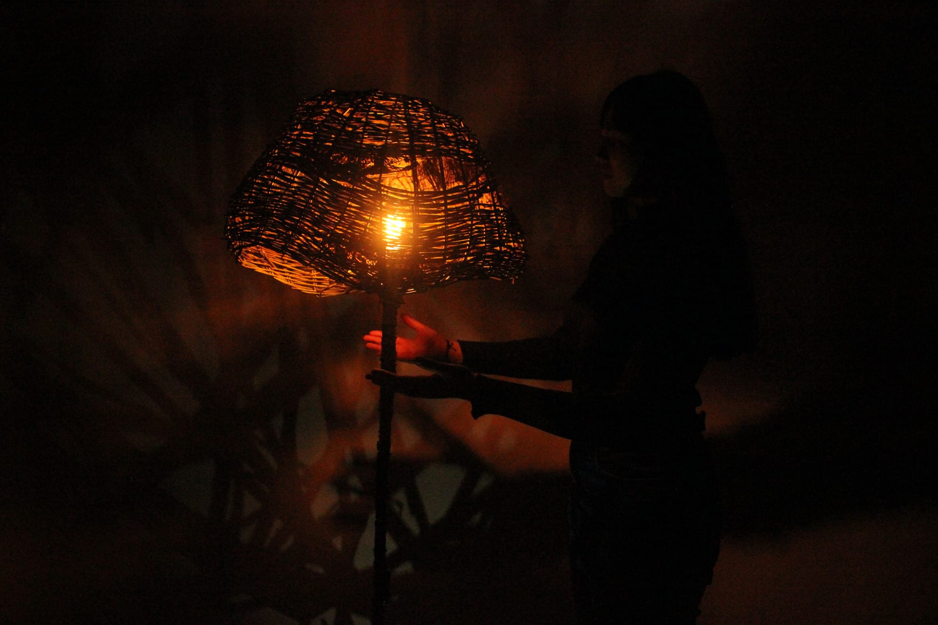 Willow woven lampshade lit from within throwing irregular shadows on wall behind with shadowy figure holding hands beneath 