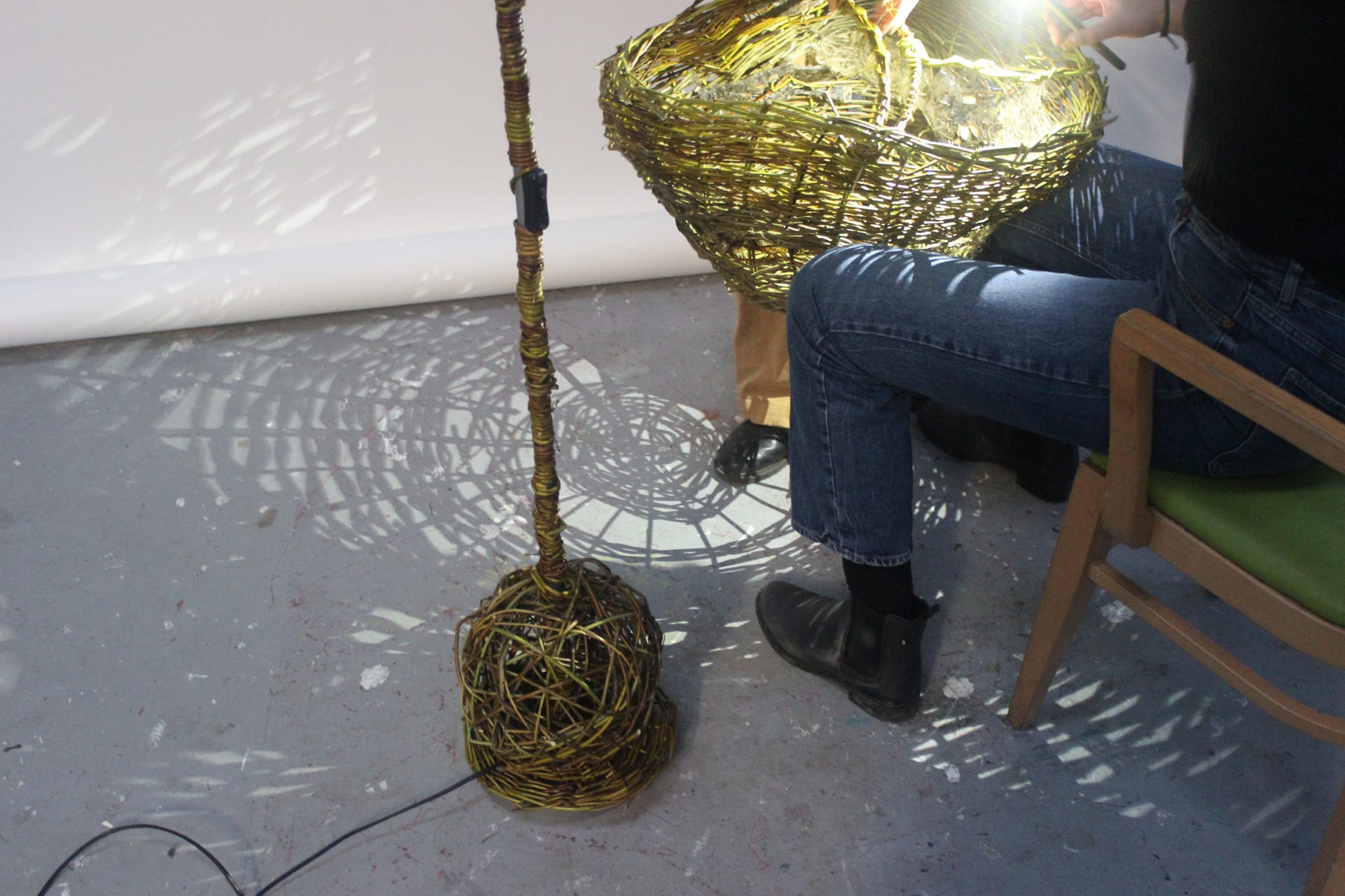 Seated woman holding lampshade shining torch into lampshade resulting in woven willow shadows on the floor and back wall