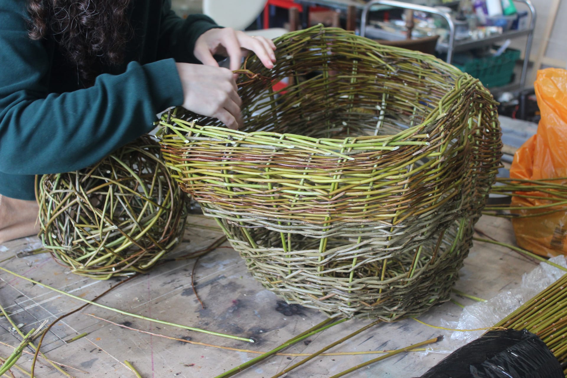 Women willow weaving lampshade in progress on a work bench, willow woven ball to the left
