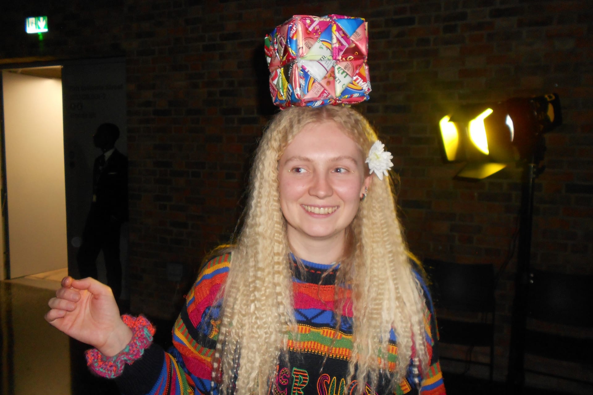 Photograph of the artist Tyler Danielle Potter with crimped blonde hair, a flower, a colourful jumper and artwork on her head.