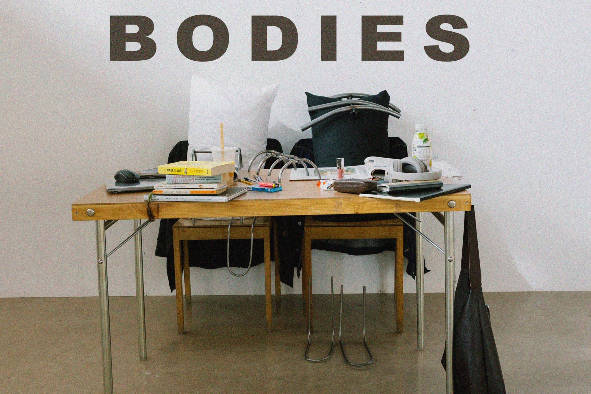 A cluttered table with various items and two pillows, metal sculptures seated on a chair. The word "BODIES" on top of the image.