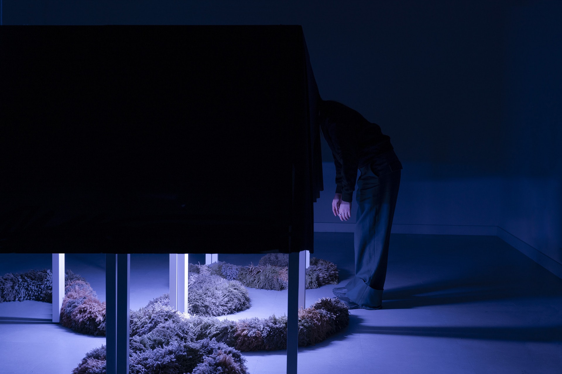 Dark room with a purple light source illuminating a central installation box, surrounded by textile art pieces.