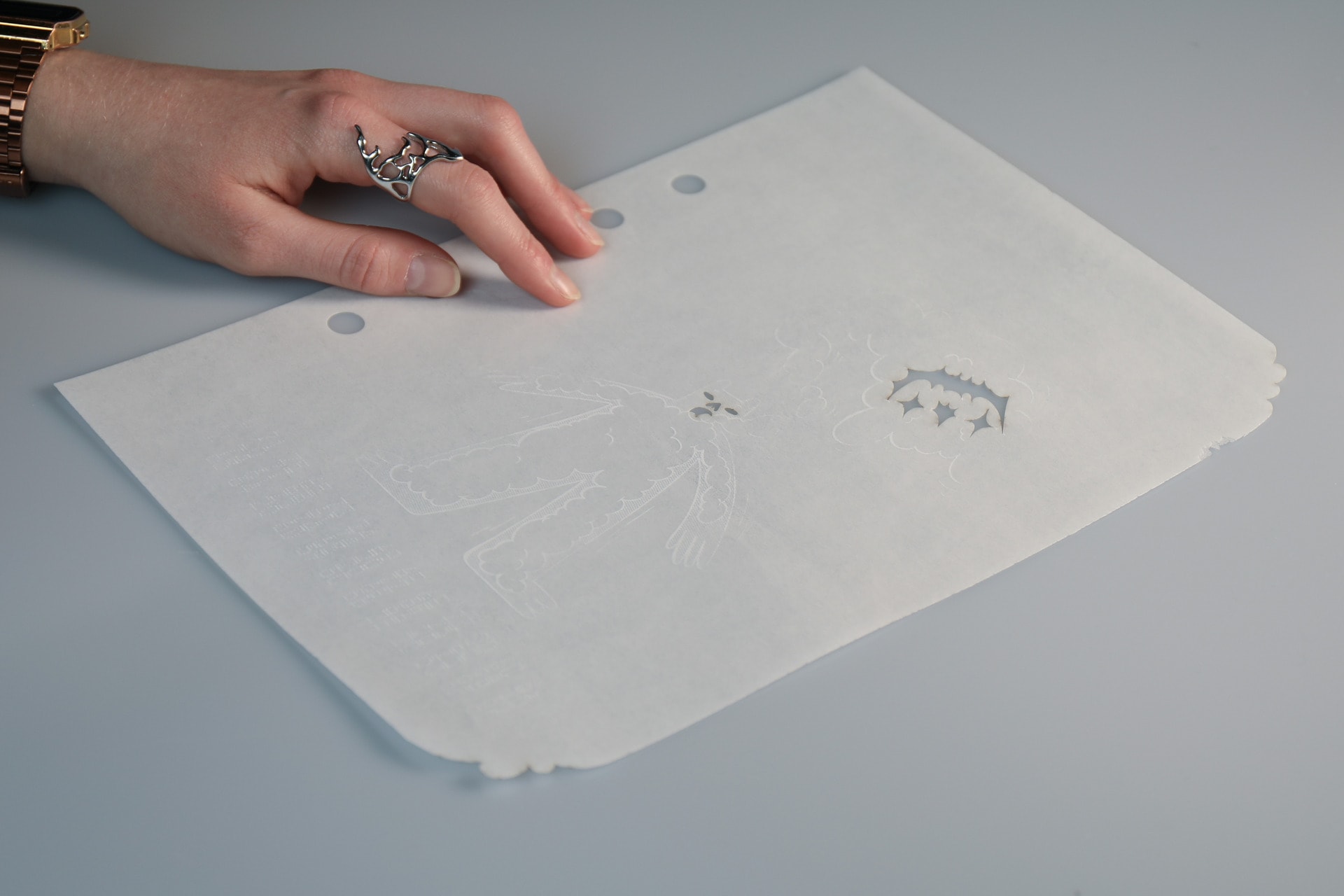 Photograph of a left hand touching the edible booklet page showing human figure with facial features cut away using the laser cutter