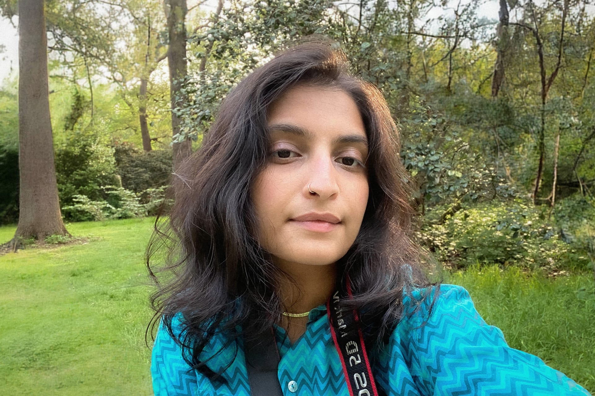 The artist in a blue shirt with a camera strap worn around her neck. The background is a lush green park in the summertime.