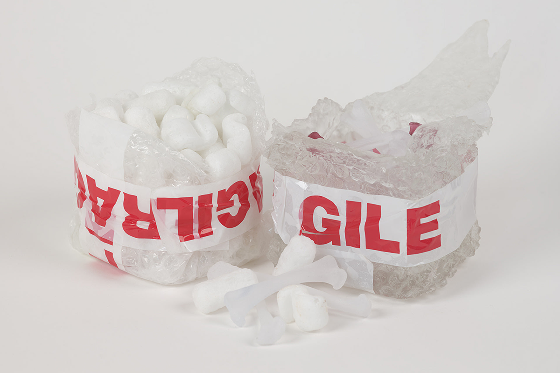 2 clear boxes with red and white fragile tape, 1 box is full of foamed glass packing peanuts, the other clear glass bones