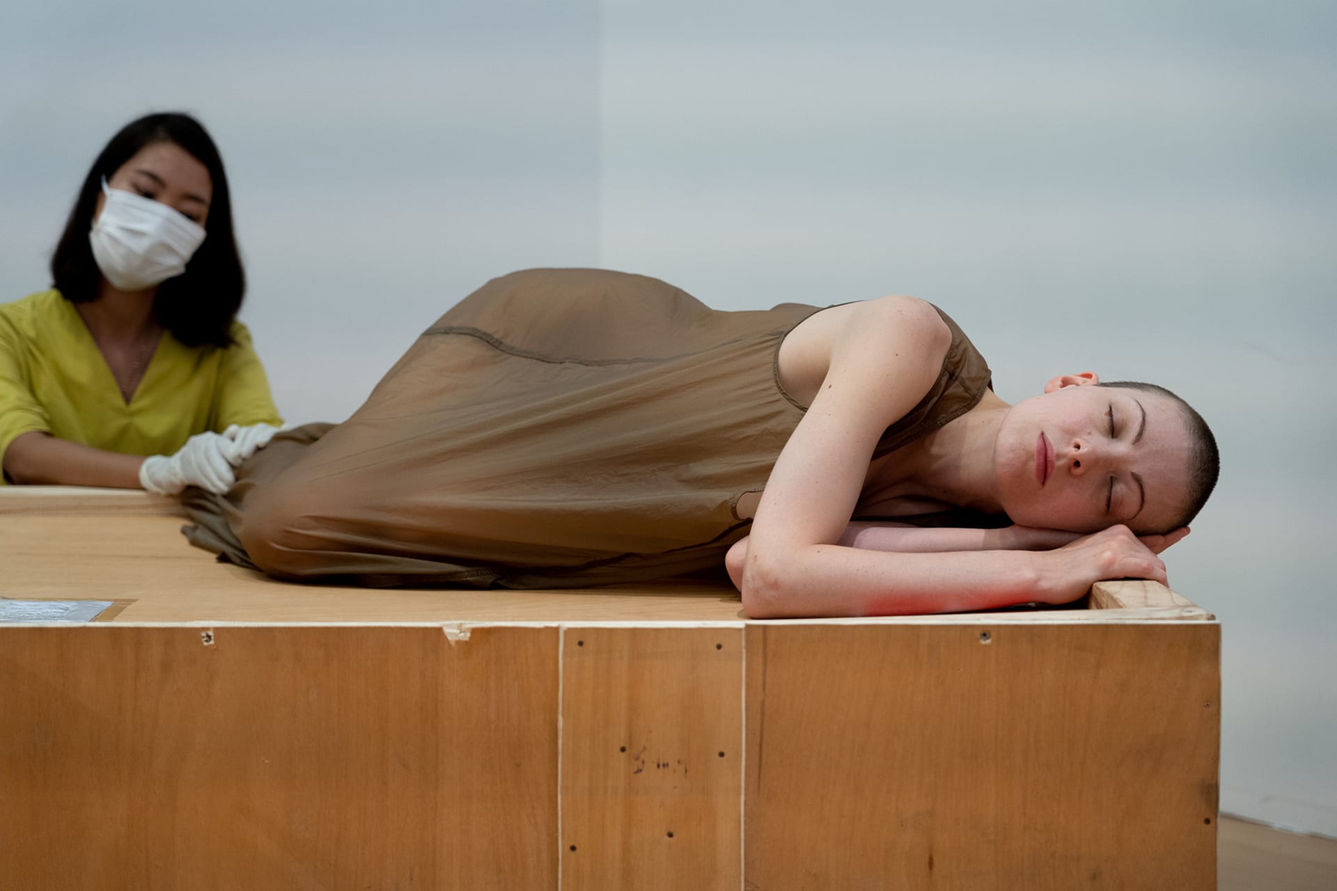 Shave-head female performance artist lying on a wooden crate with her eyes closed. A girl wearing latex gloves touches her. 