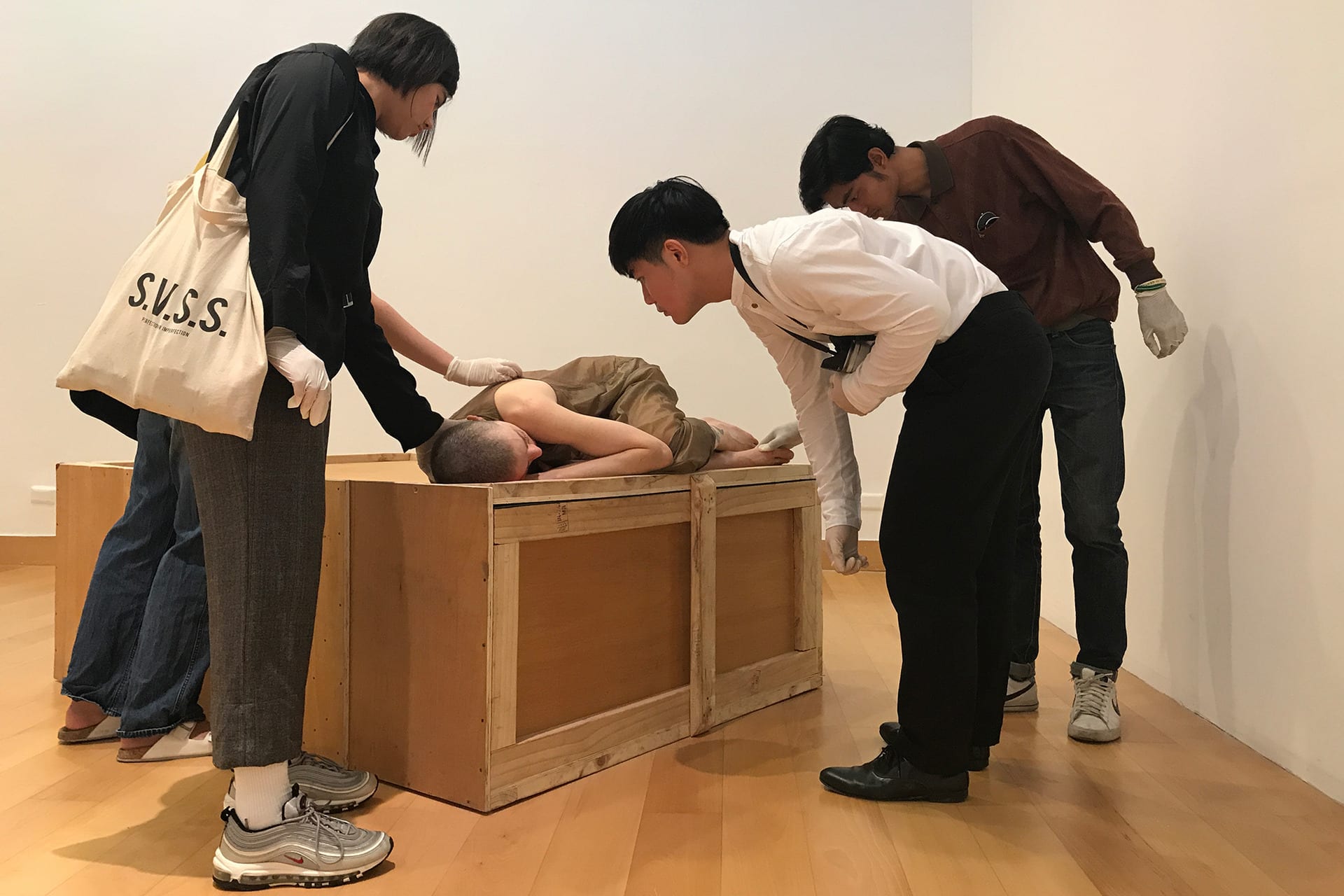 Shaved-head female performer in a semi-transparent dress, lying on a crate. A group of spectators touching her with latex gloves