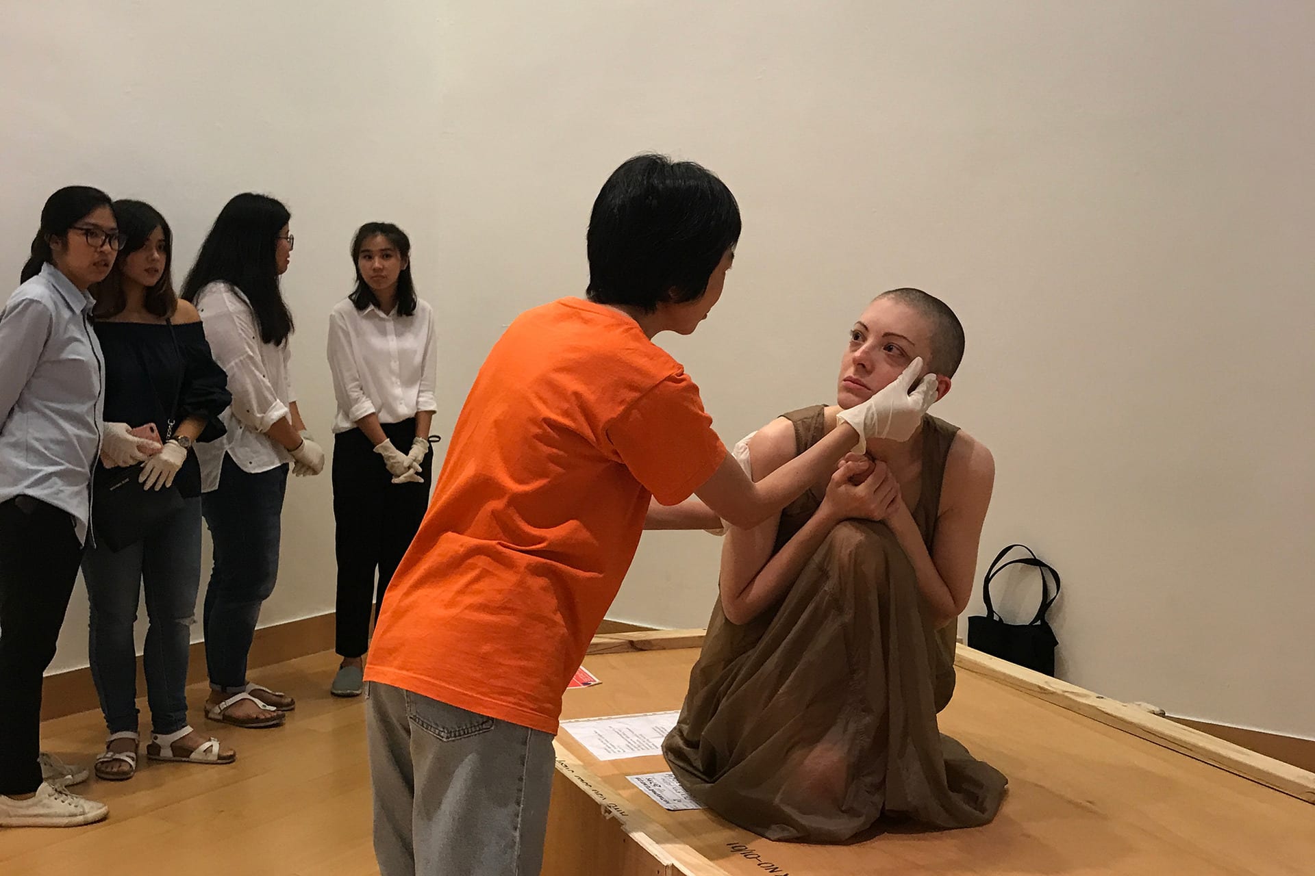 Shaved-head female performer in a semi-transparent dress, kneeling on a crate. A spectator touching her head with latex gloves