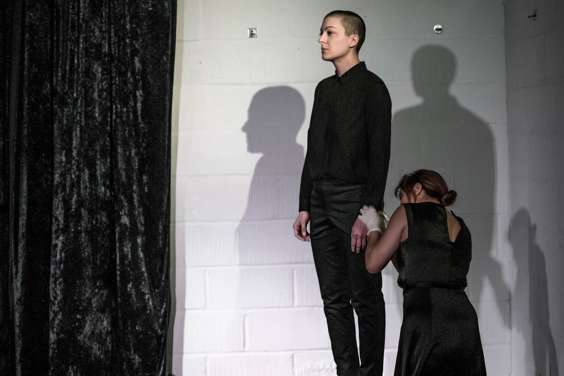 Shaved-head female performer in black shirt and pants, standing on a crate. A female spectator touching her with latex gloves. 