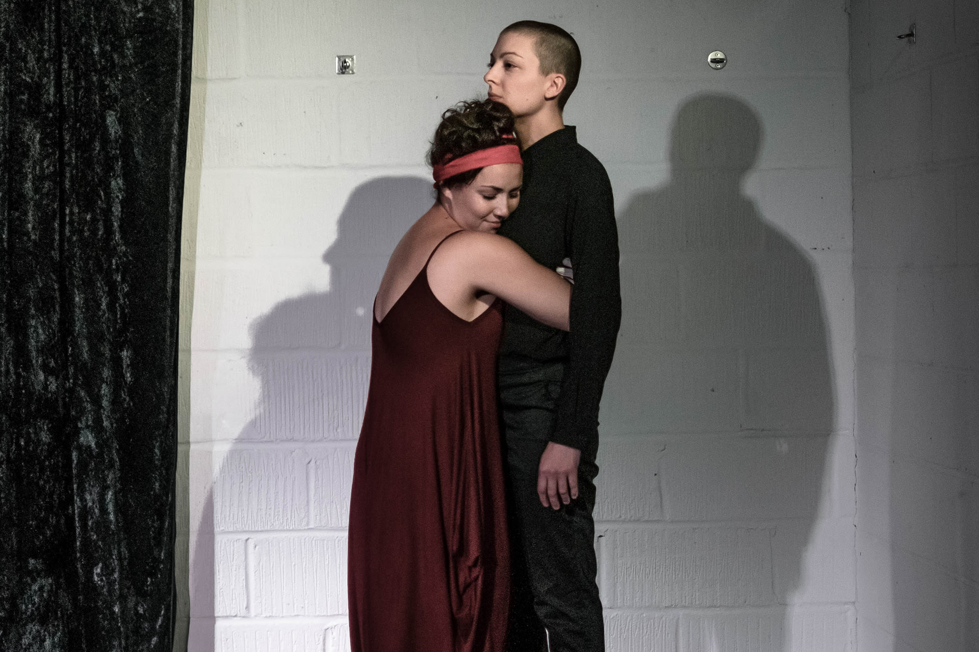 Shaved-head female performer in black shirt and pants, standing on a crate. A female spectator hugging her. 