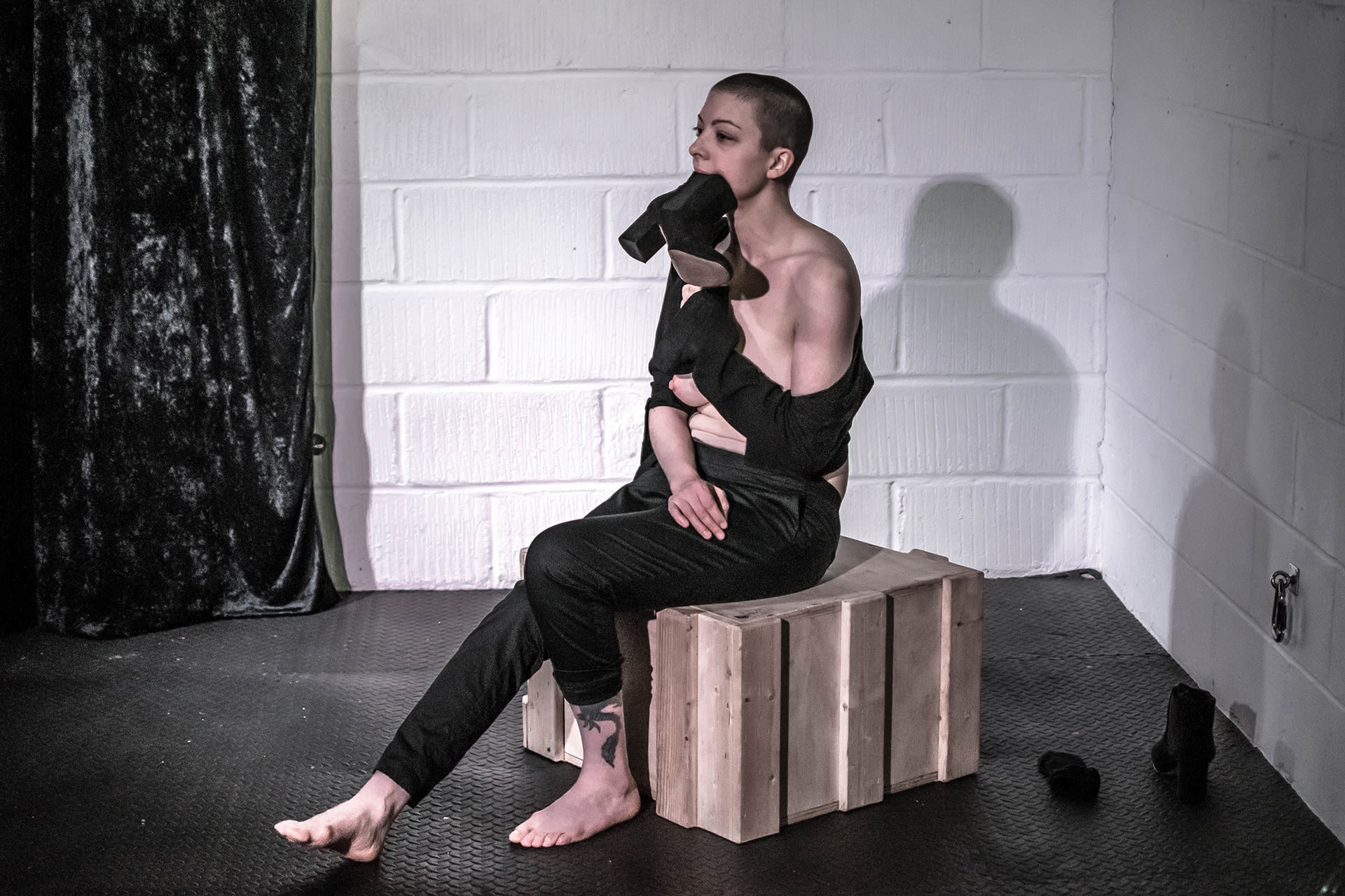 Shaved-head female performer in black shirt and pants, sitting on a crate, half-naked, with a show in her mouth. 