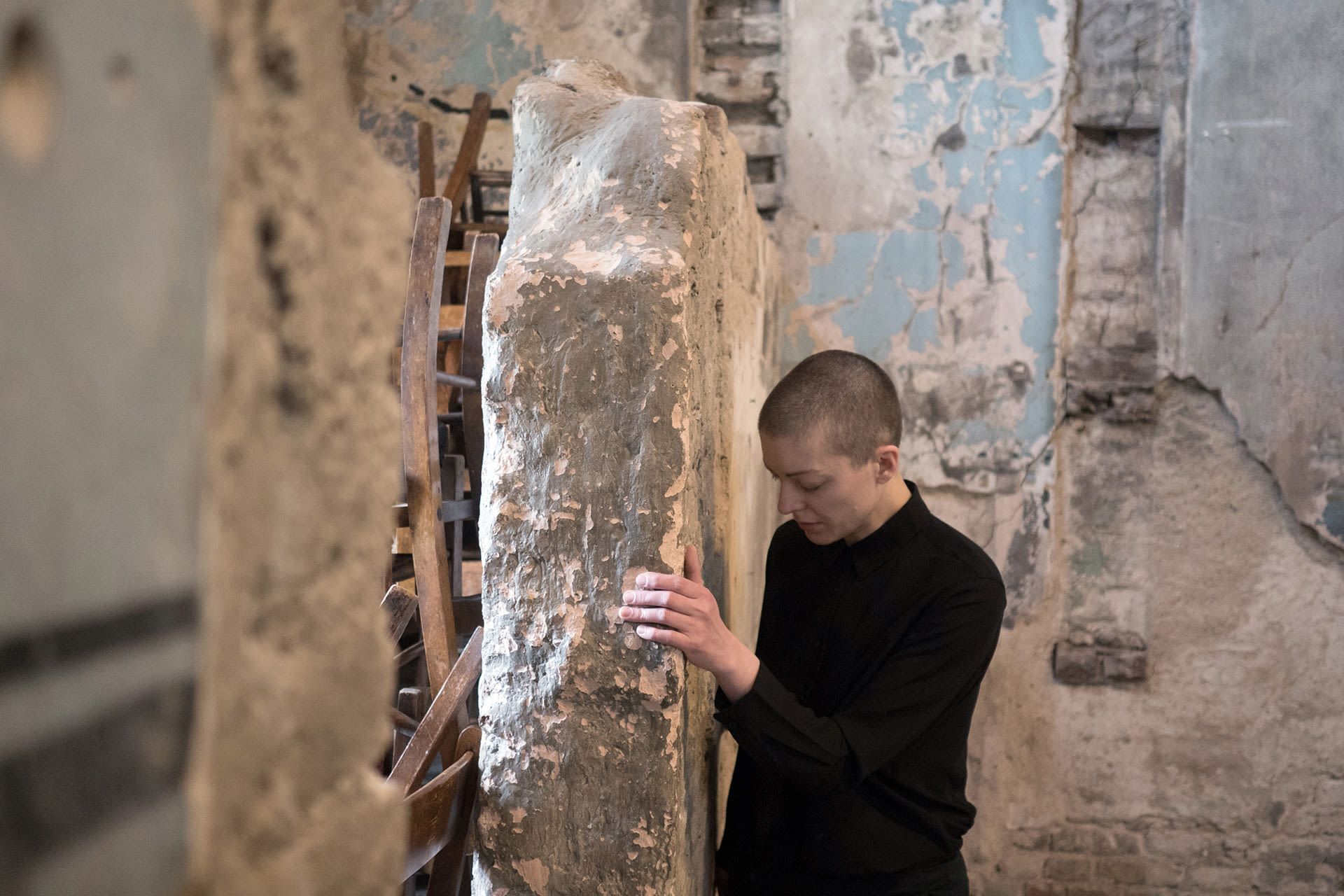 Shaved-head female performer in black shirt and pants, walking while facing and touching an old chapel's wall. 