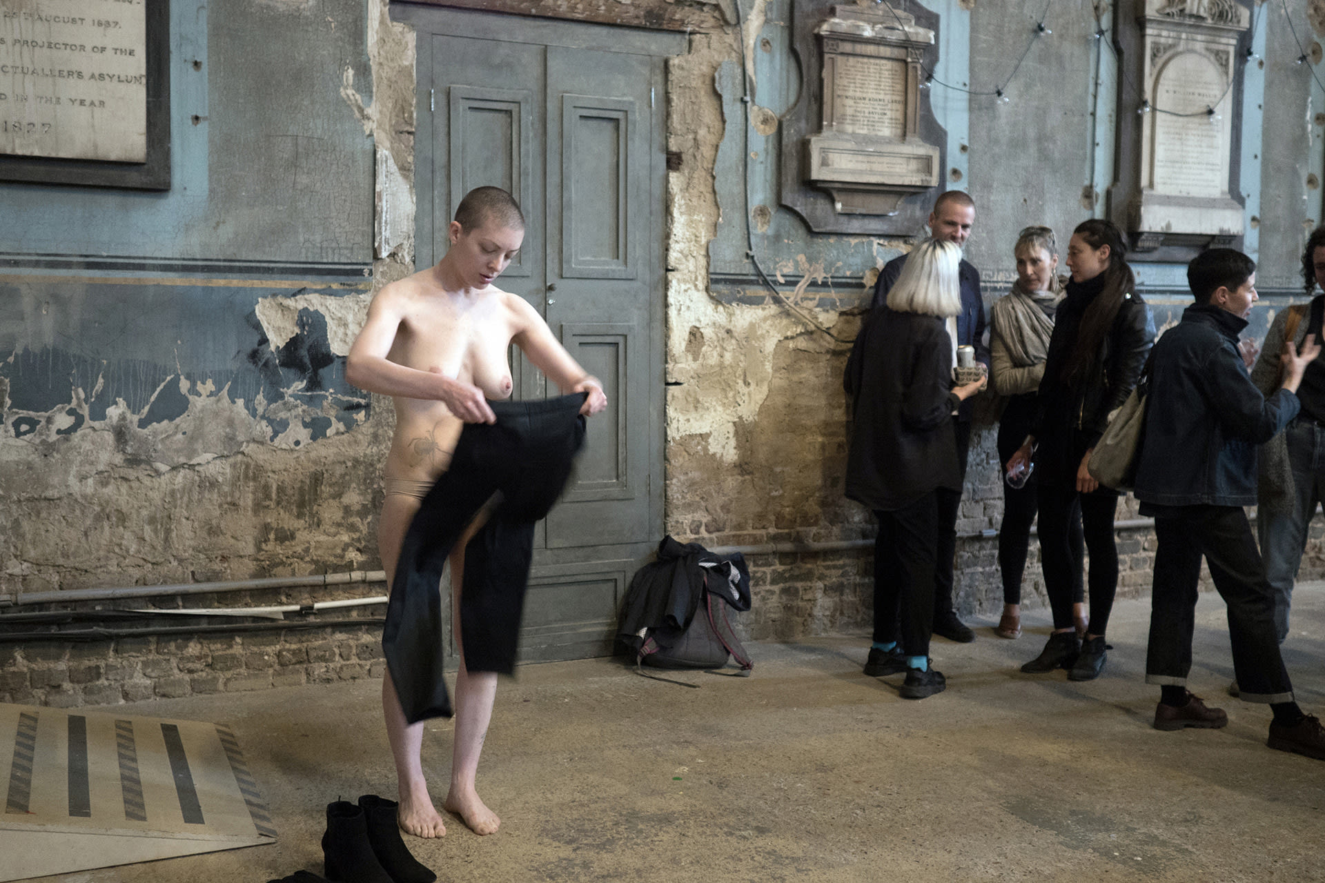 Shaved-head female performer in black shirt and pants, taking off her clothes. 