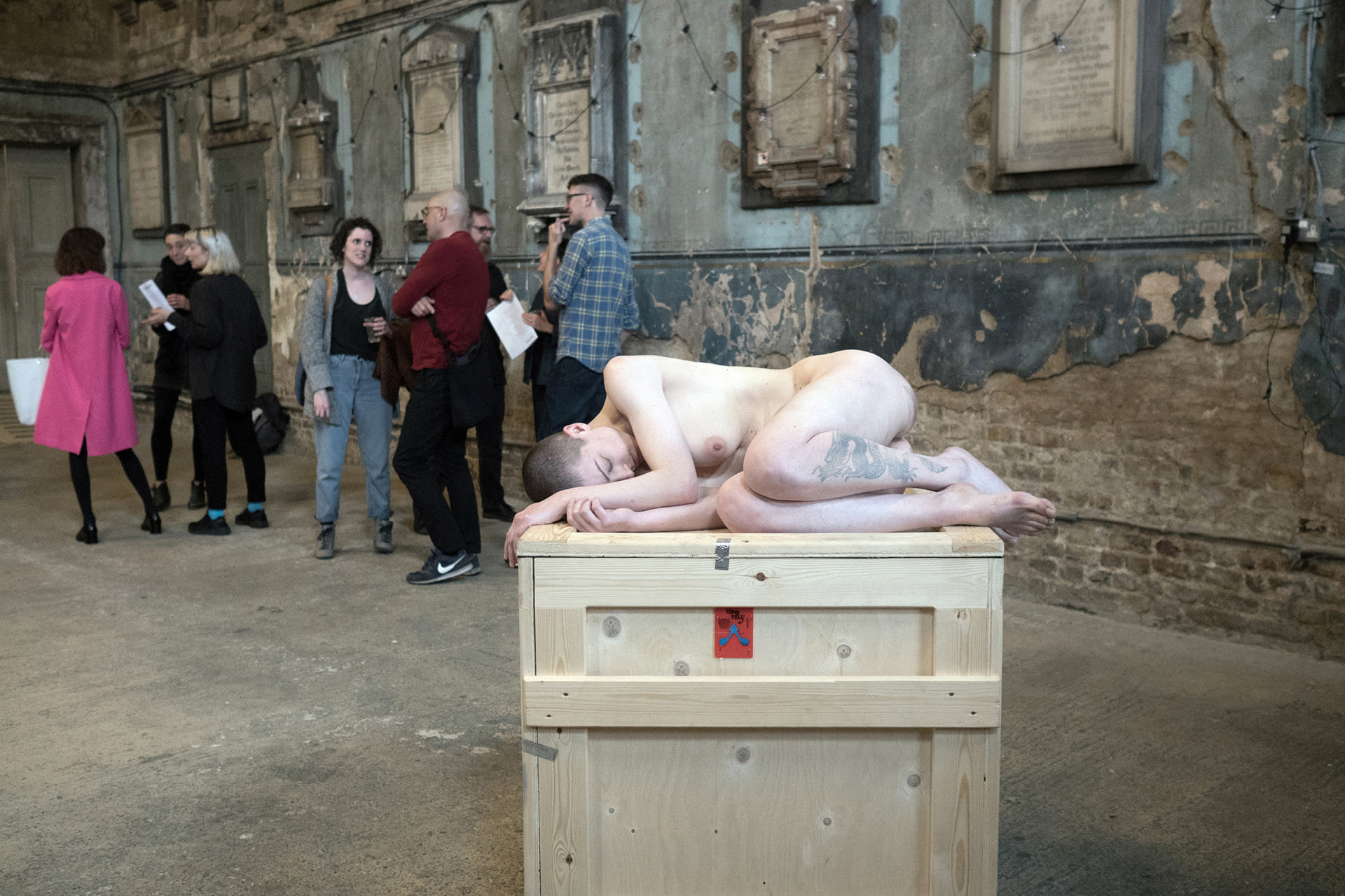 Shaved-head female performer lying naked on a crate in a former Asylum's chapel. 