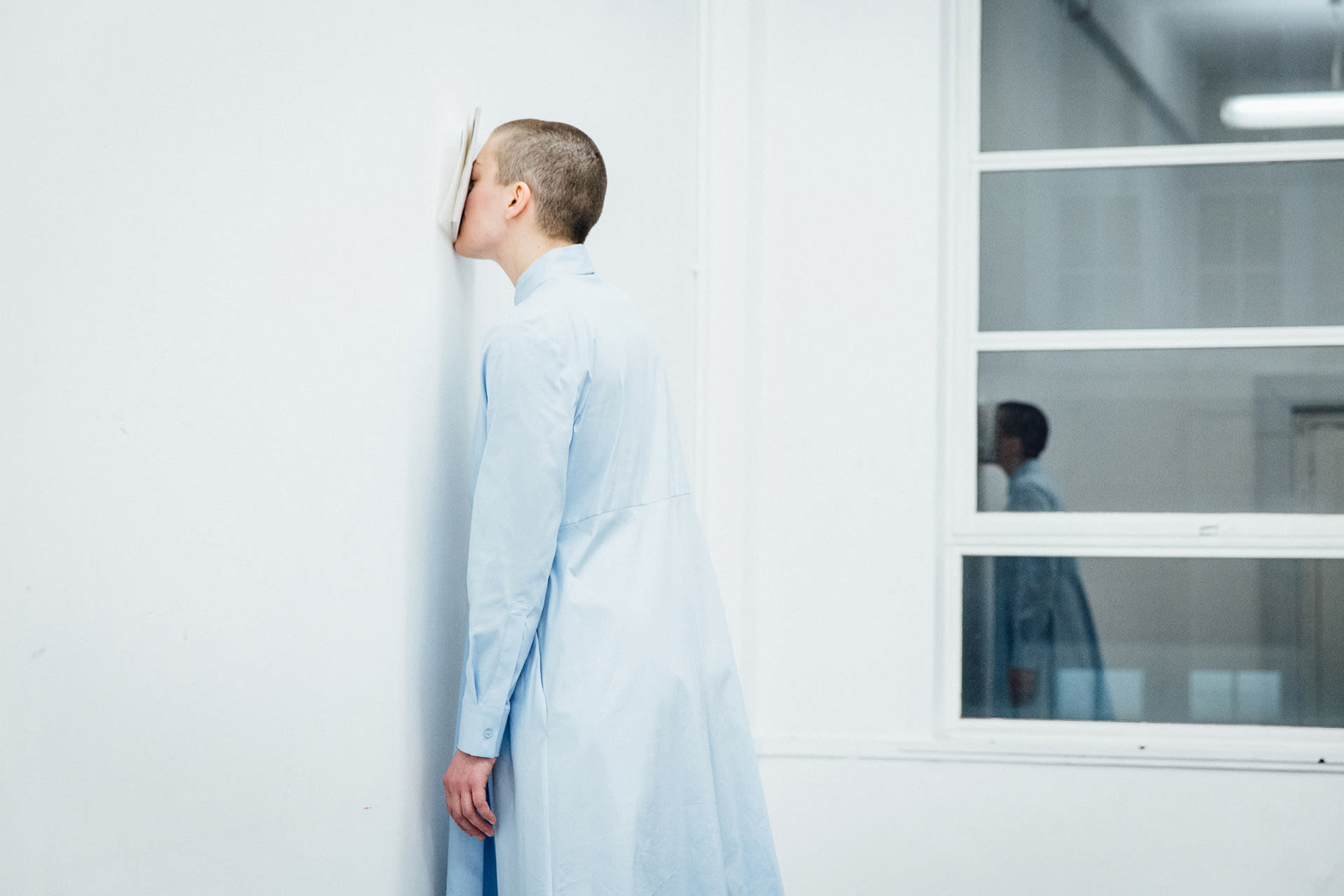 Shaved-head female performer in light blue dress, standing on her toes, with her face pressing a book against a white wall. 