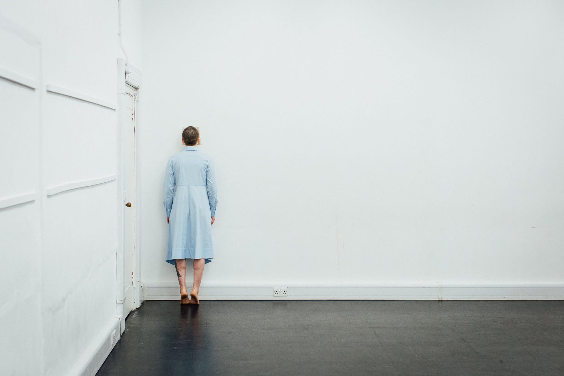 Shaved-head female performer in light blue dress, standing on her toes, with her face pressing a book against a white wall. 