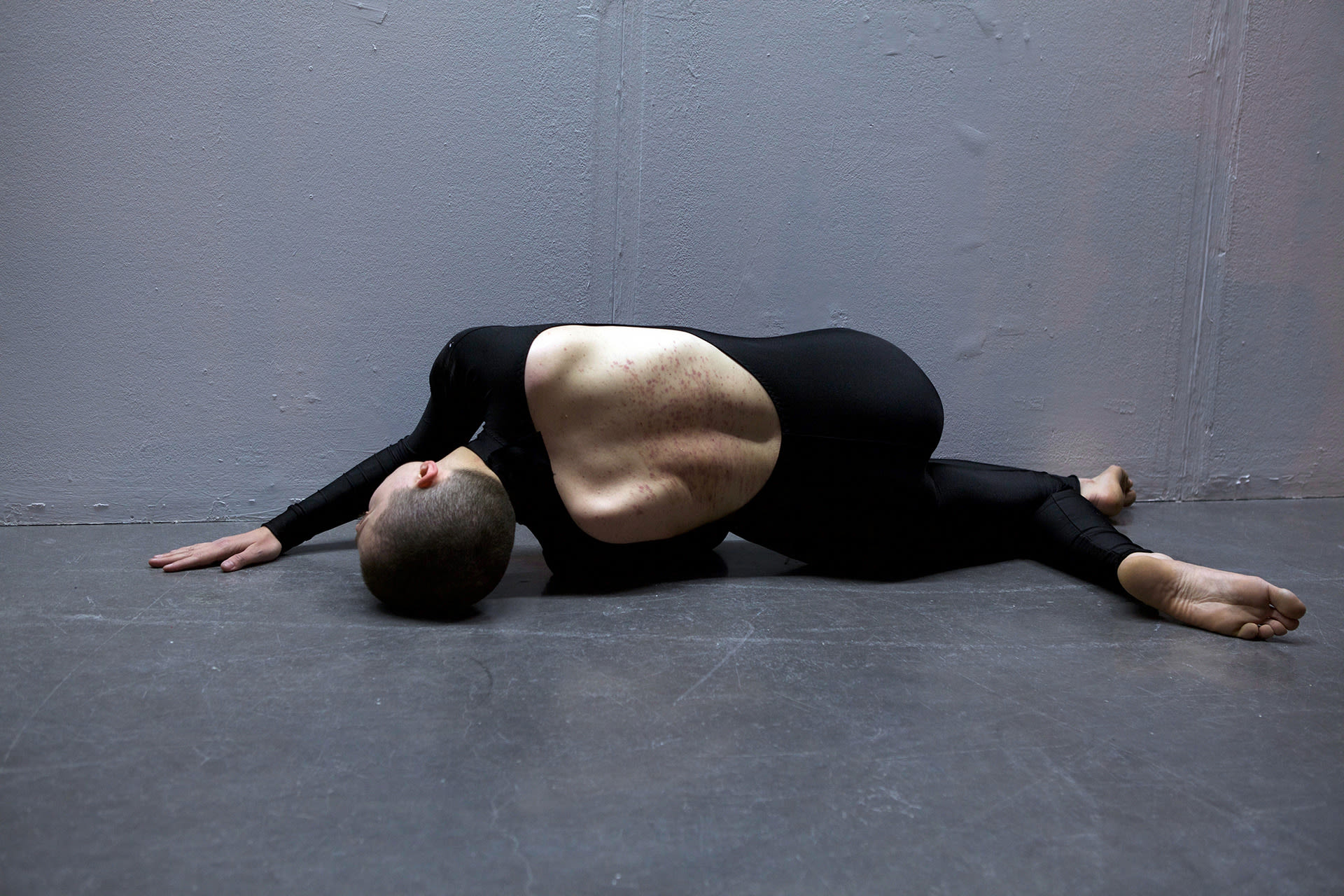 Shaved-head female performer crawling on the floor, wearing a black full body leotard with the back open. Her back bears marks. 