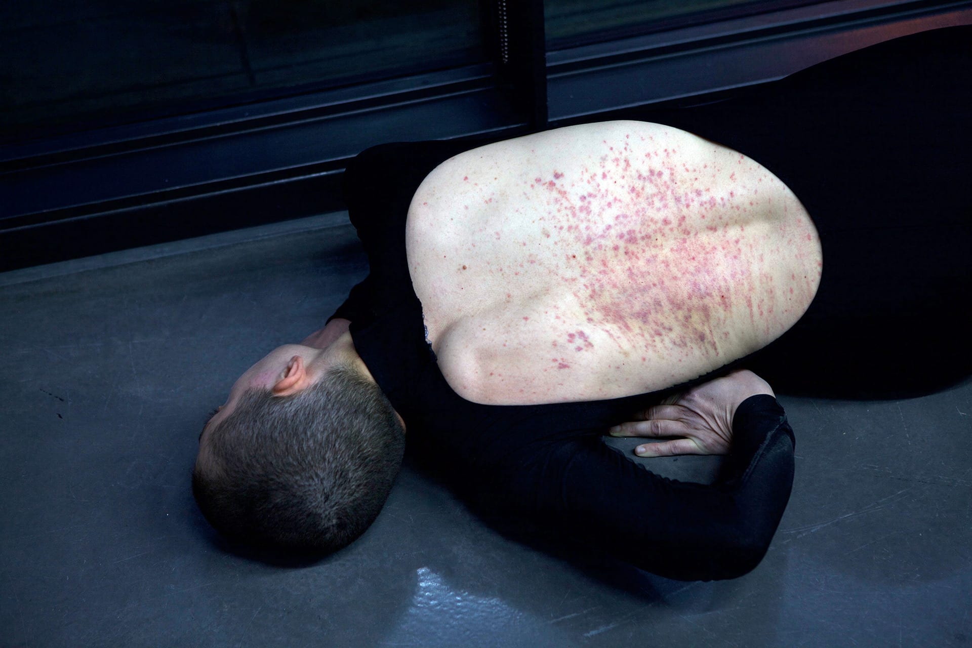 Shaved-head female performer crawling on the floor, wearing a black full body leotard with the back open. Her back bears marks. 