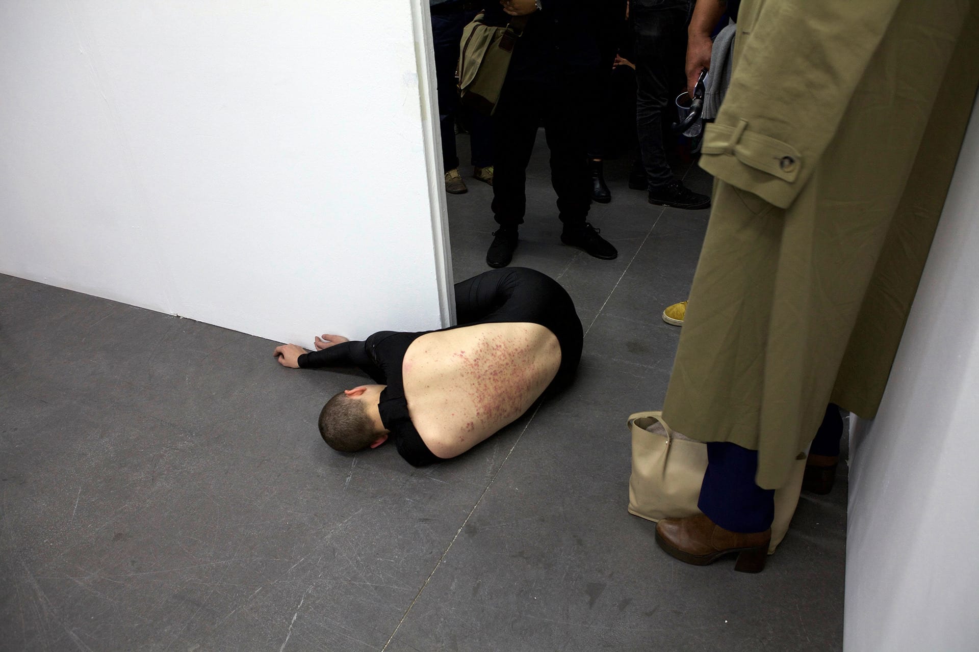 Shaved-head female performer crawling on the floor, wearing a black full body leotard with the back open. Her back bears marks. 