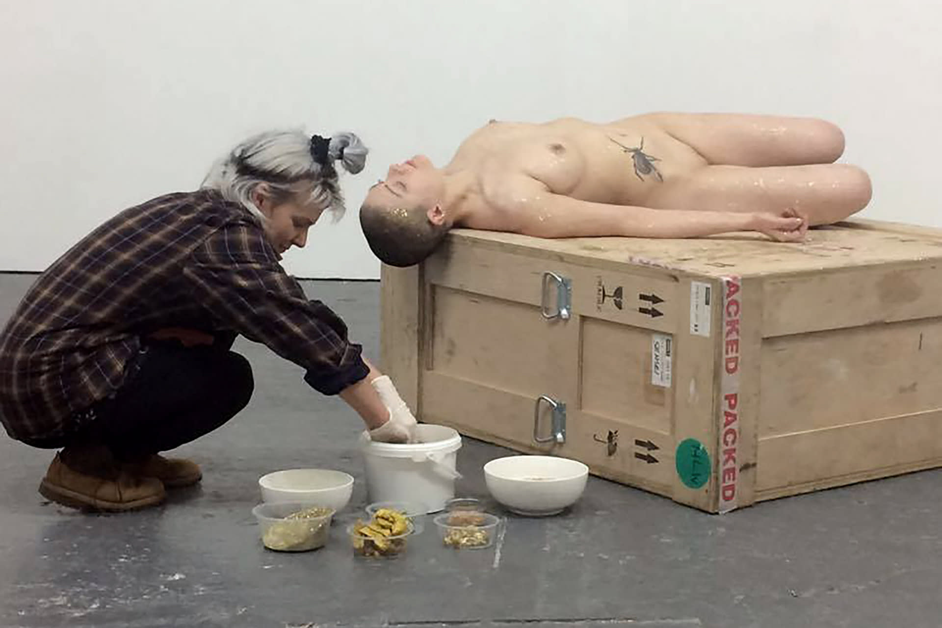 Shaved-head female performer lying on a crate, having a female visitor applying honey and glitter on her body. 