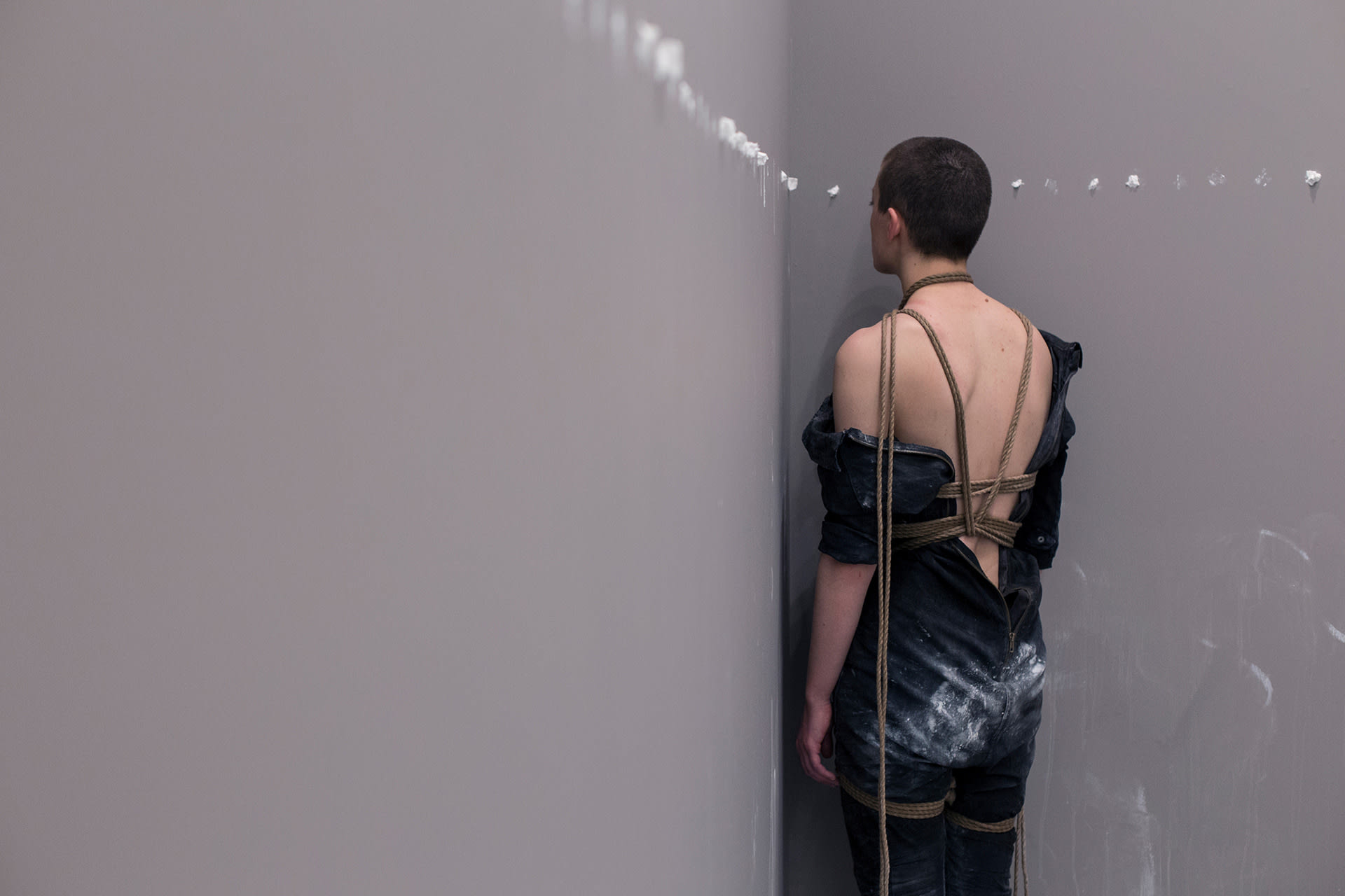 Shaved-head female performer facing the corner of a room, dressed in a worker's outfit, with her back exposed, in bondage. 