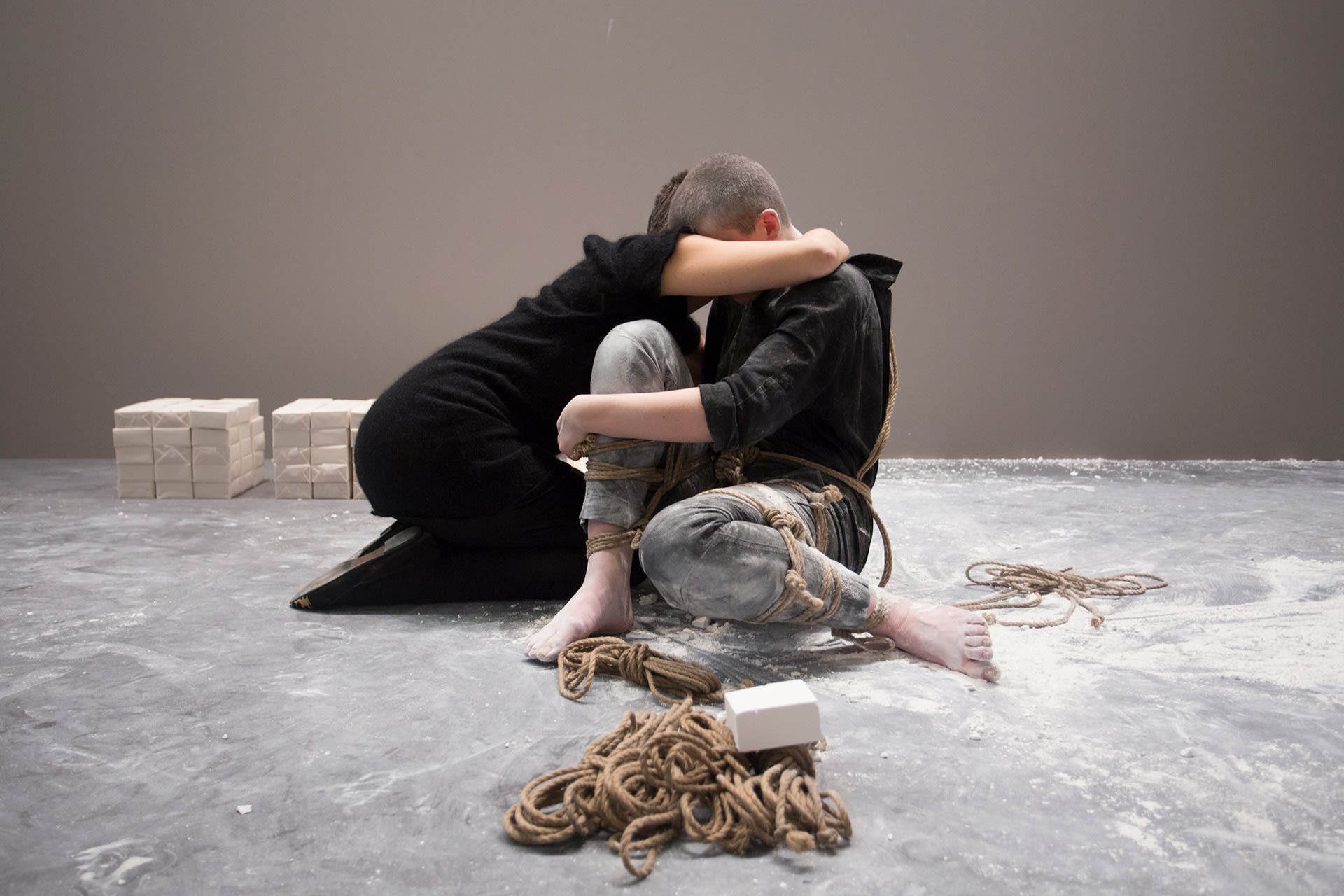 Shaved-head female performer in a worker's uniform, in bondage, sitting on the floor and being hugged by a visitor.